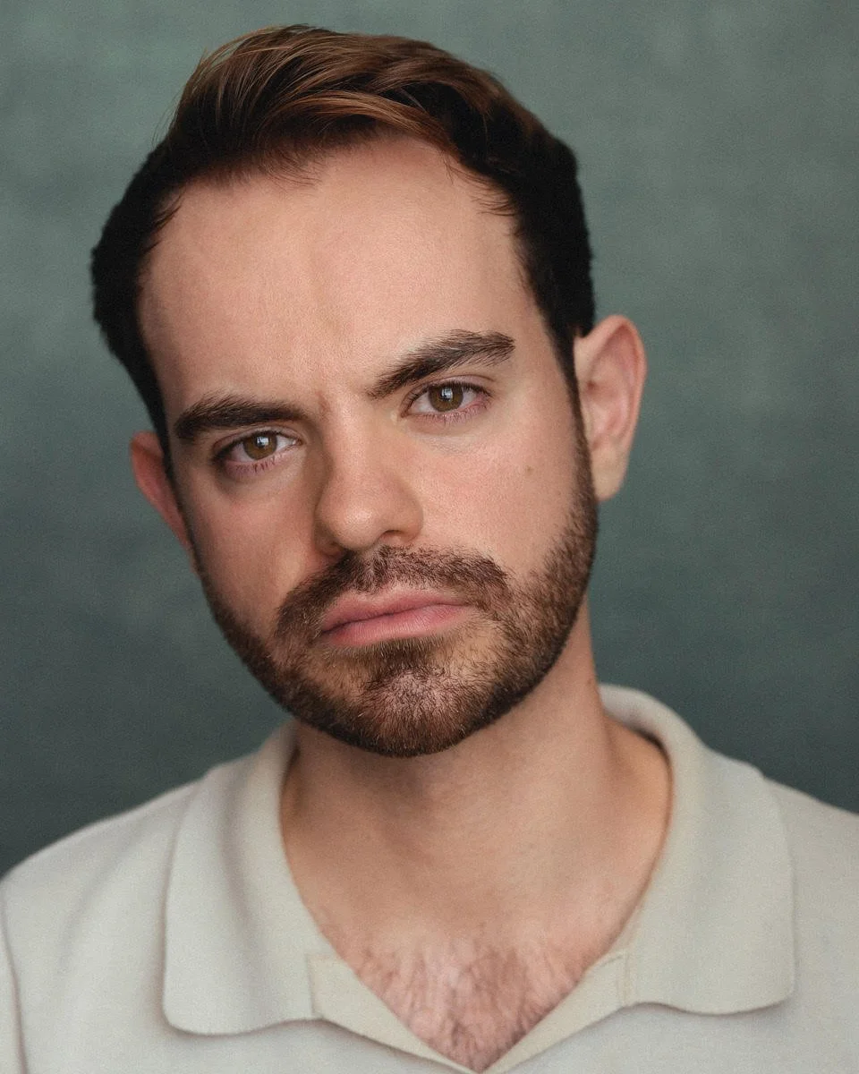 Close-up portrait of a young man with brown hair, hazel eyes, and a beard, wearing a light-colored shirt against a muted green background.
