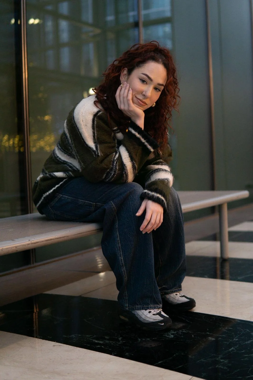A young woman with curly red hair sitting on a bench indoors, resting her chin on her hand, wearing a striped sweater, jeans, and sneakers.