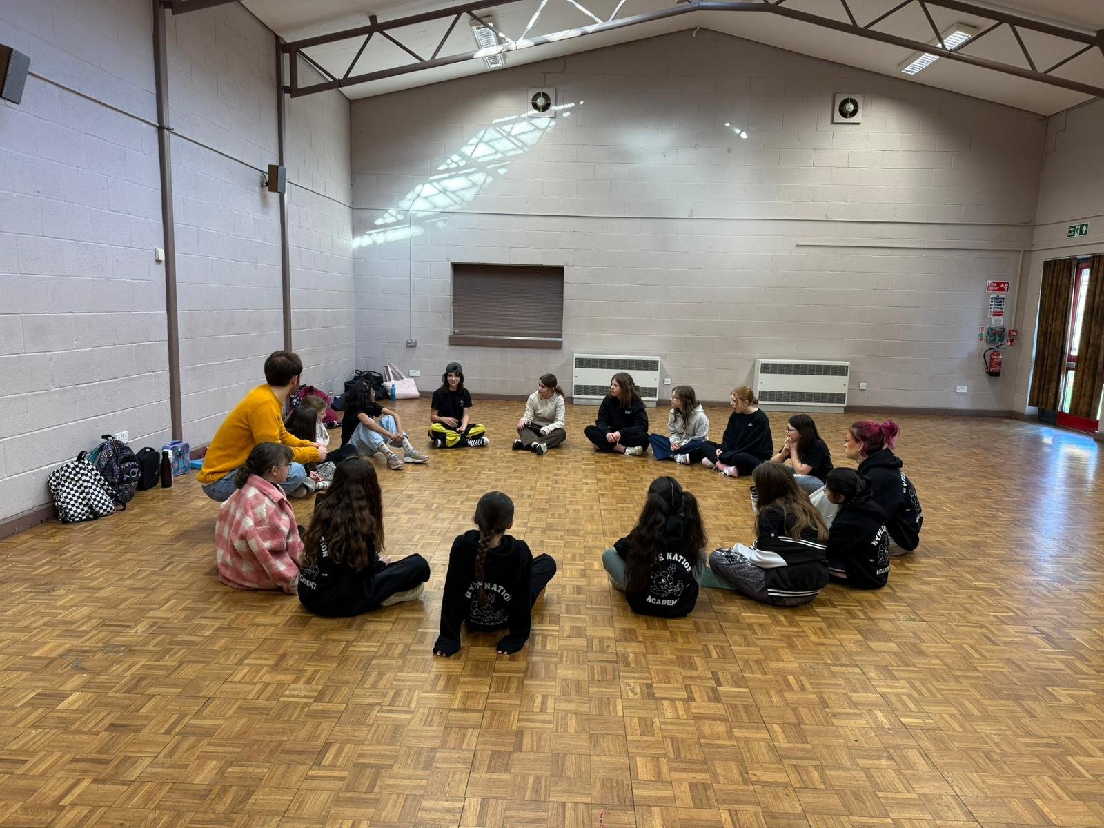 Group of young girls and women sitting in a circle on a wooden floor inside a gymnasium or community center, with backpacks and bags beside them.