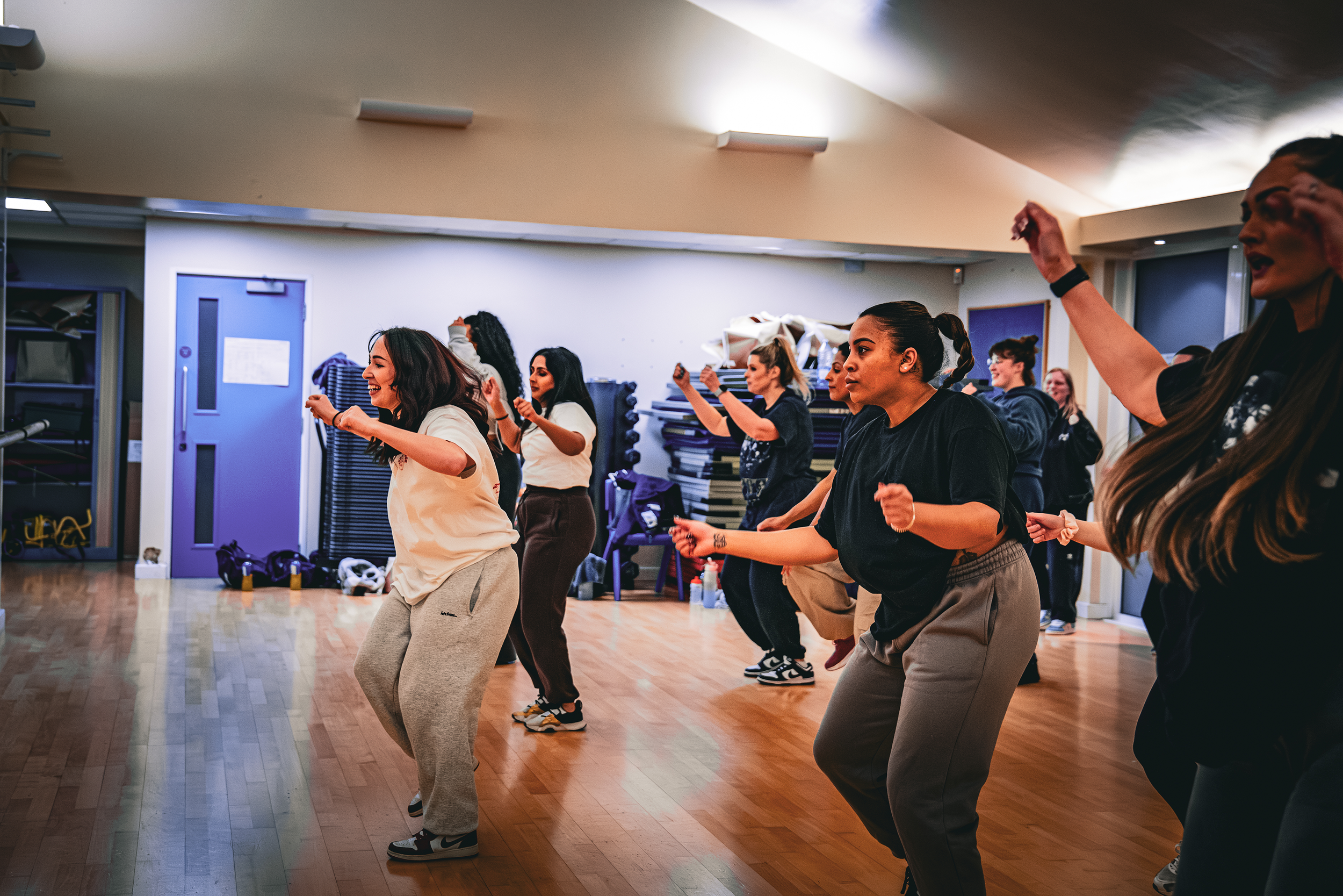 A group of women participating in a dance or fitness class in a studio, moving energetically with smiles and focused expressions.