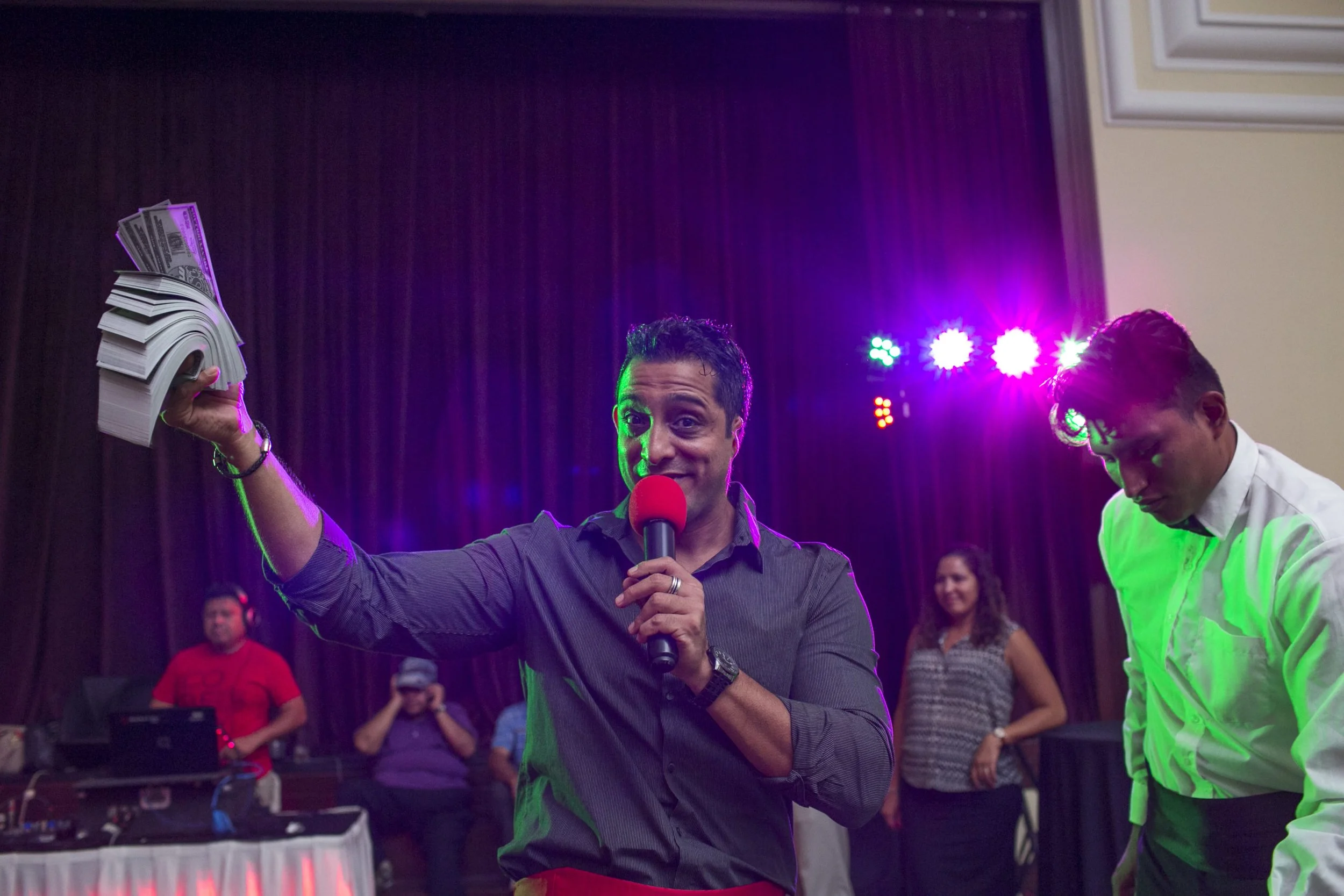 A man holding a microphone and a stack of dollar bills on stage with purple and green lighting, with several people observing in the background.