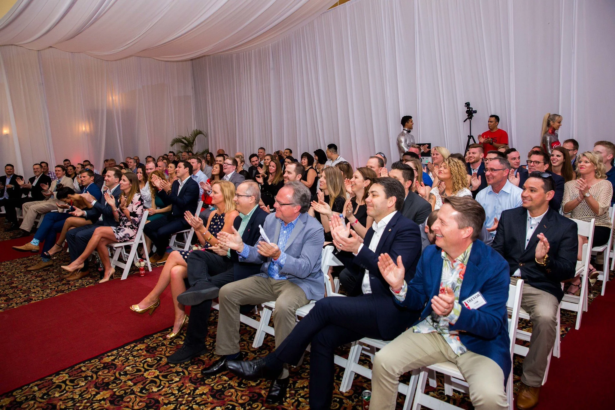 A large group of people seated and clapping at an indoor event with white draped walls and carpeted floor.