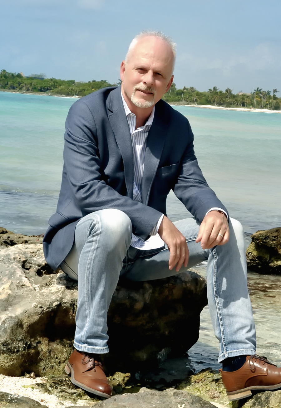 A man in a blue blazer and gray jeans sitting on rocks by the beach with turquoise water and palm trees in the background.