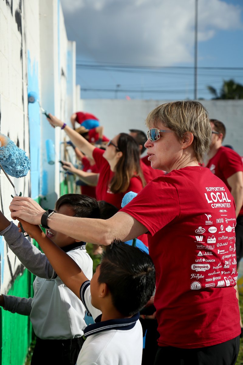 People, including children and adults, painting a mural on a wall outdoors. They are wearing red shirts, and the scene appears to be part of a community service event.