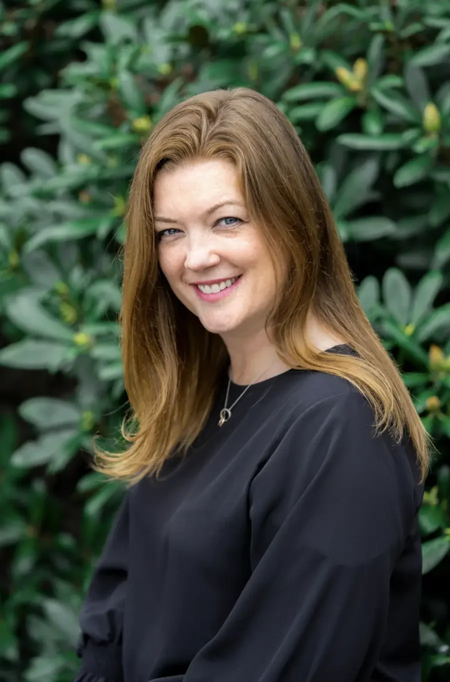 A woman with long, reddish-brown hair and blue eyes, smiling at the camera, standing outdoors in front of green leafy plants, wearing a black top and a silver necklace.