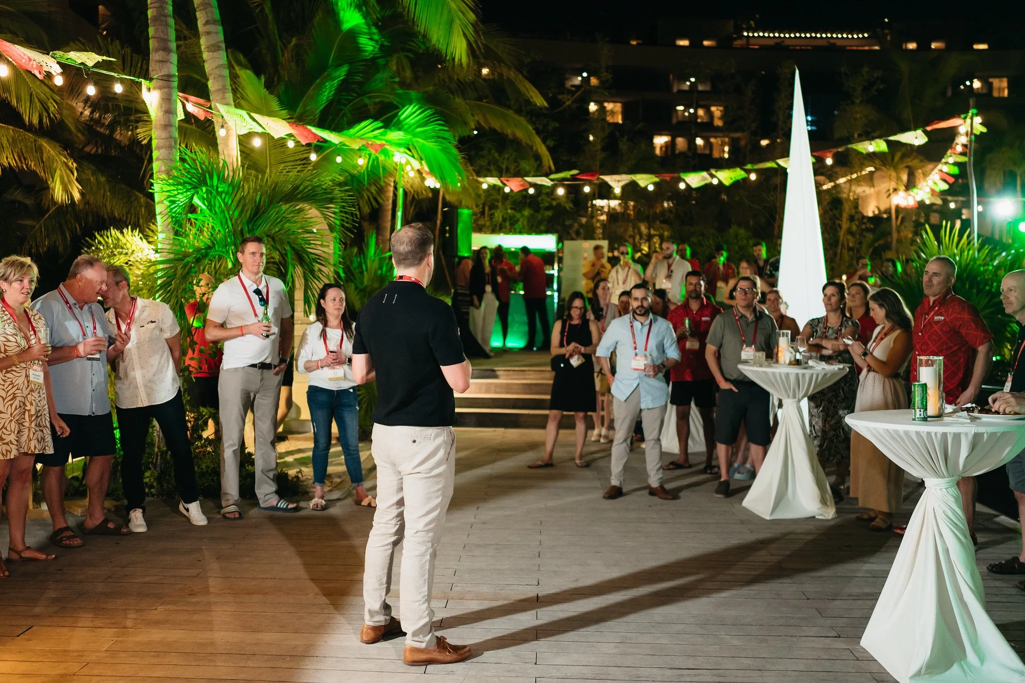 People gathered outdoors at a nighttime event with string lights, palm trees, and tropical decor, including a large white cone-shaped structure and tables with drinks.