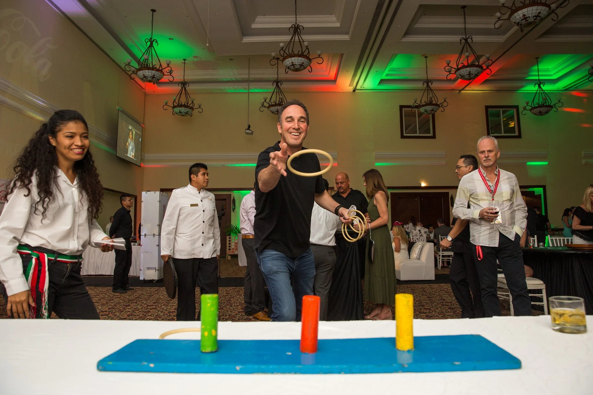 People playing a game where they throw rings onto colorful pegs on a table at an indoor event, with a man smiling and rotating rings in his hand, surrounded by others watching.