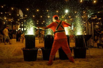 A street performer dressed as a firefighter creating sparks with metal tools at night, with a crowd watching in the background.