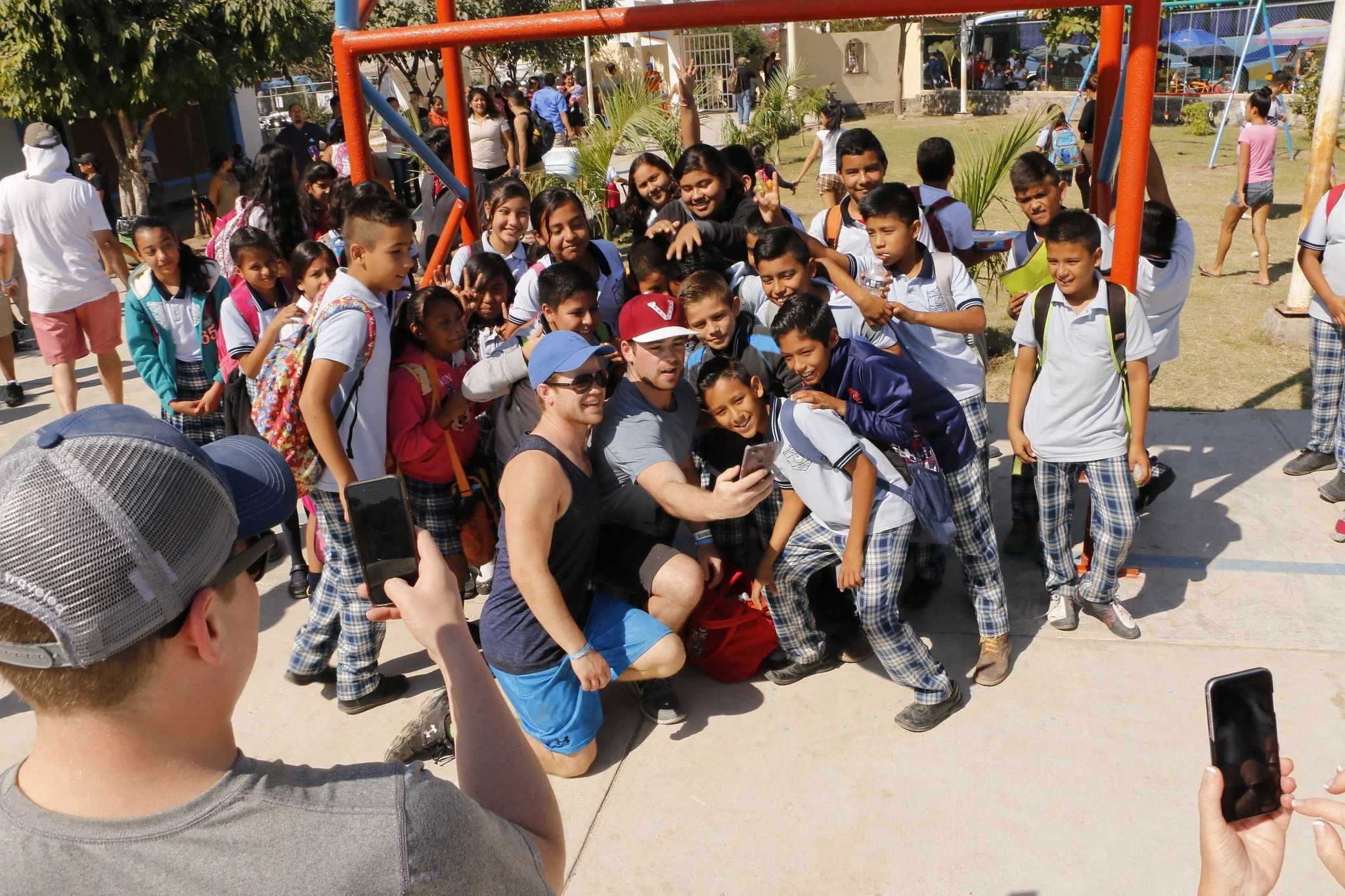 A group of school children and two adult men taking a selfie at a park or schoolyard with a playground and other children in the background.