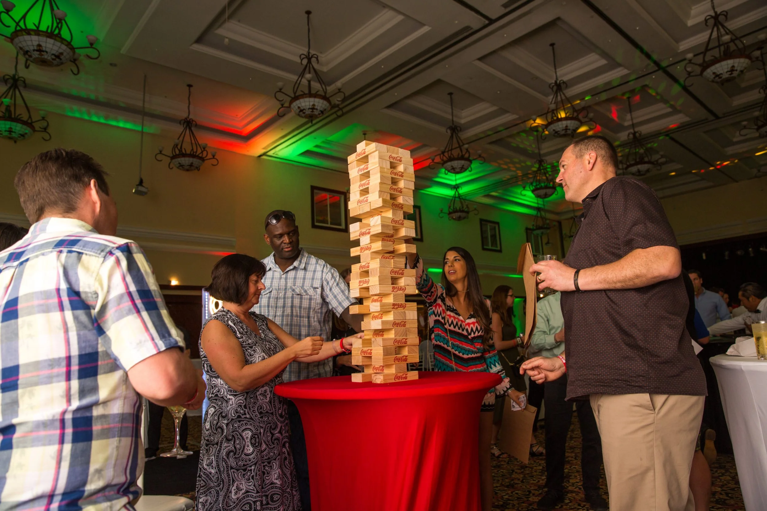People gathered around a large Jenga tower made of Coca-Cola branded wooden blocks at a social event or party in a banquet hall with colorful lighting.