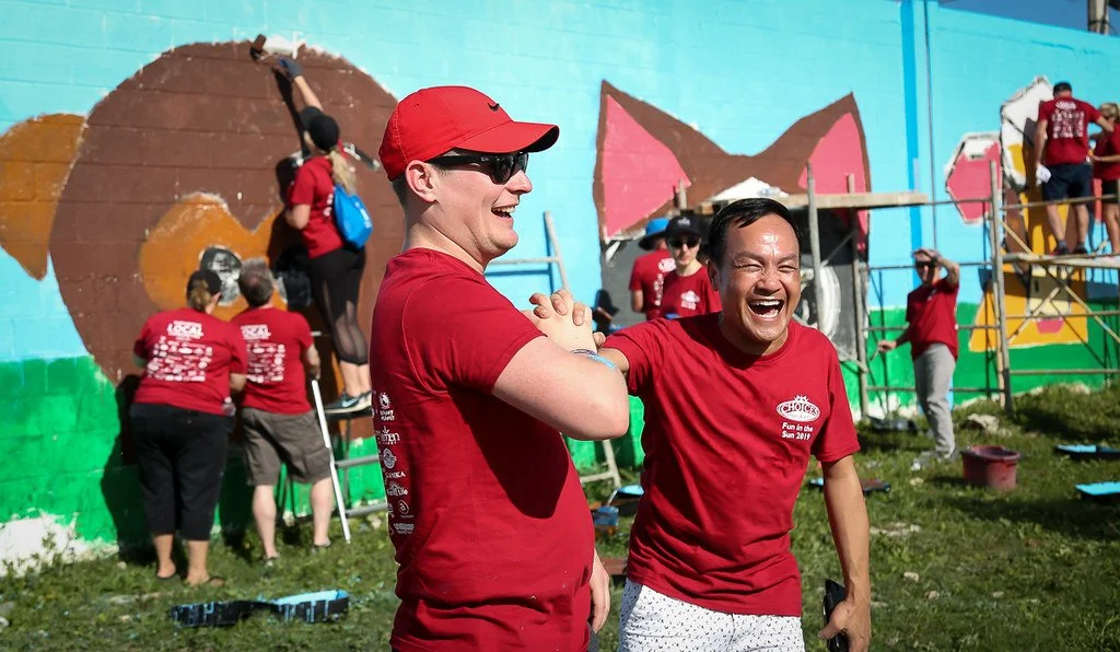 Two women wearing red shirts and sunglasses smiling and shaking hands, in front of a colorful mural being painted by volunteers in red shirts.