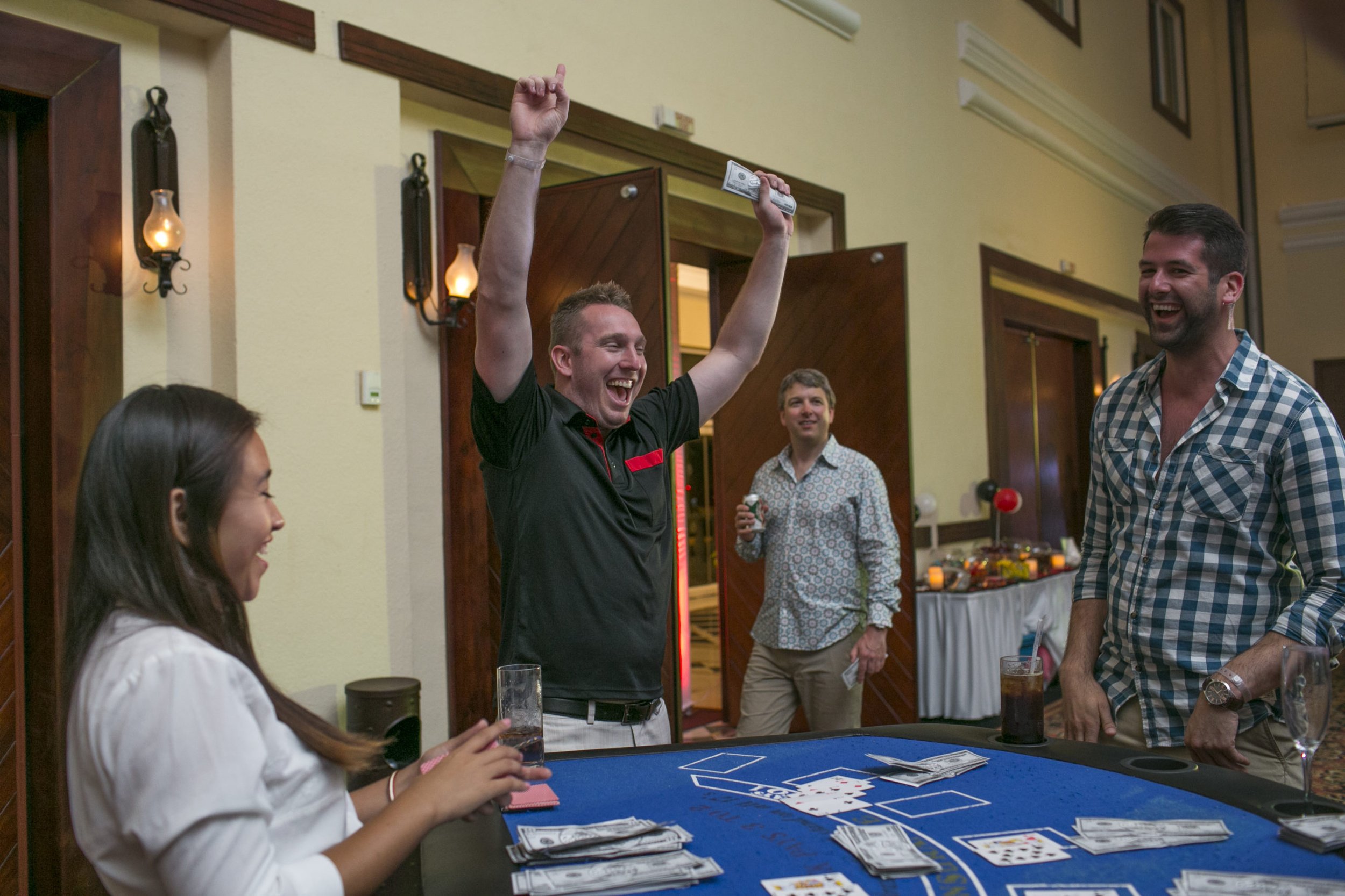 A group of people celebrating at a party, with one man holding cash in the air and smiling, while others are gathered around a table playing cards and holding drinks.