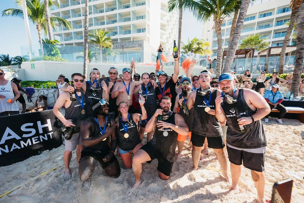 A group of men and women on a sandy beach celebrating with medals and drinks, with high-rise buildings and palm trees in the background.
