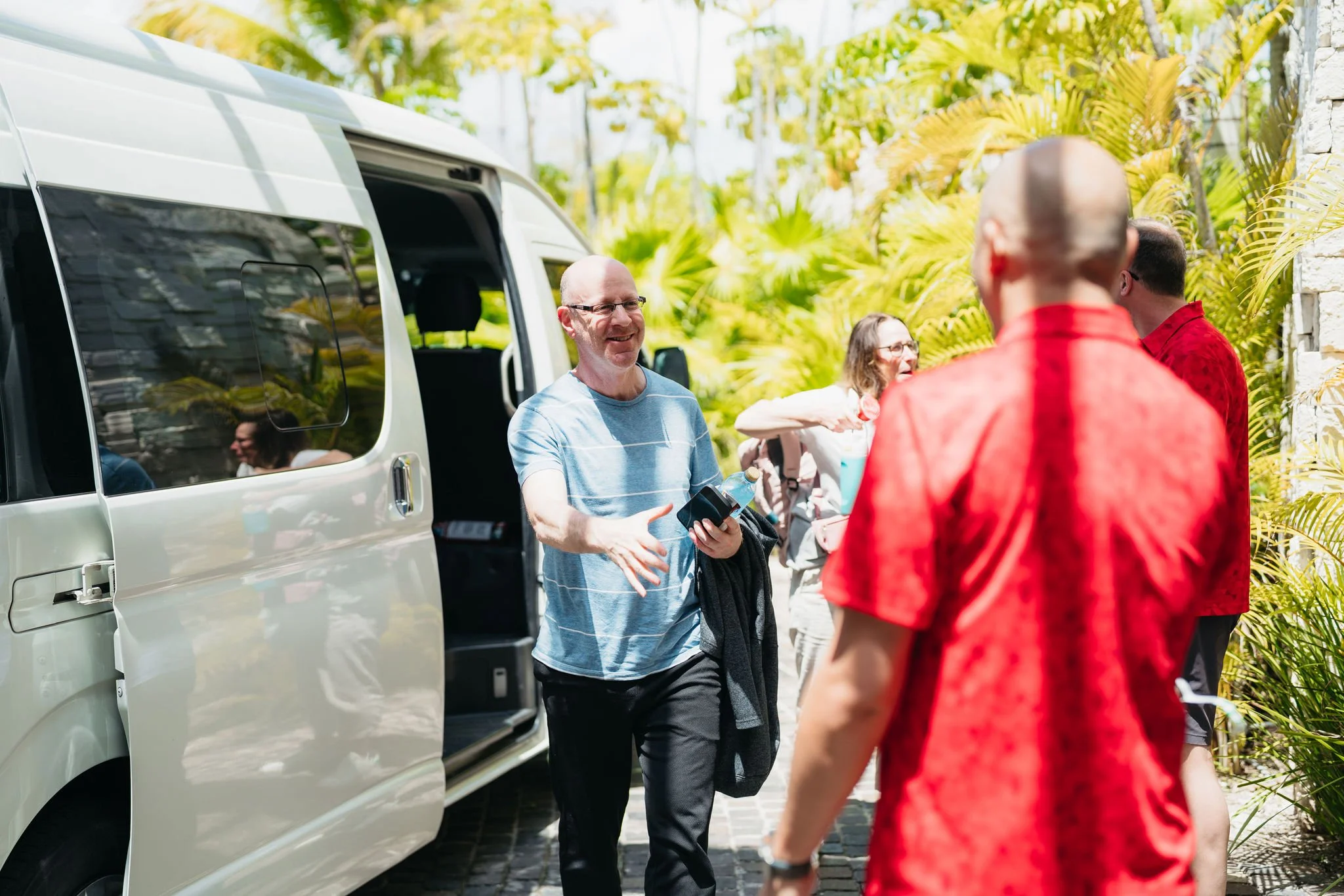 People standing outside near a white van, engaging in conversation on a sunny day with lush green foliage in the background.