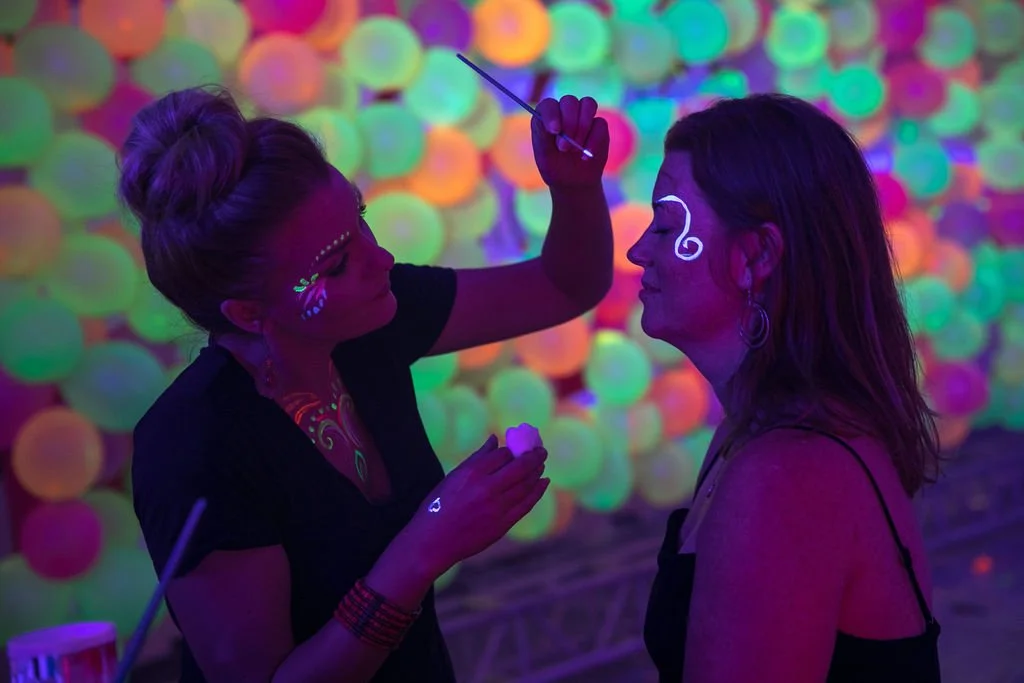 A woman is painting neon white designs on another woman's face in a glow-in-the-dark room with colorful neon balls in the background.