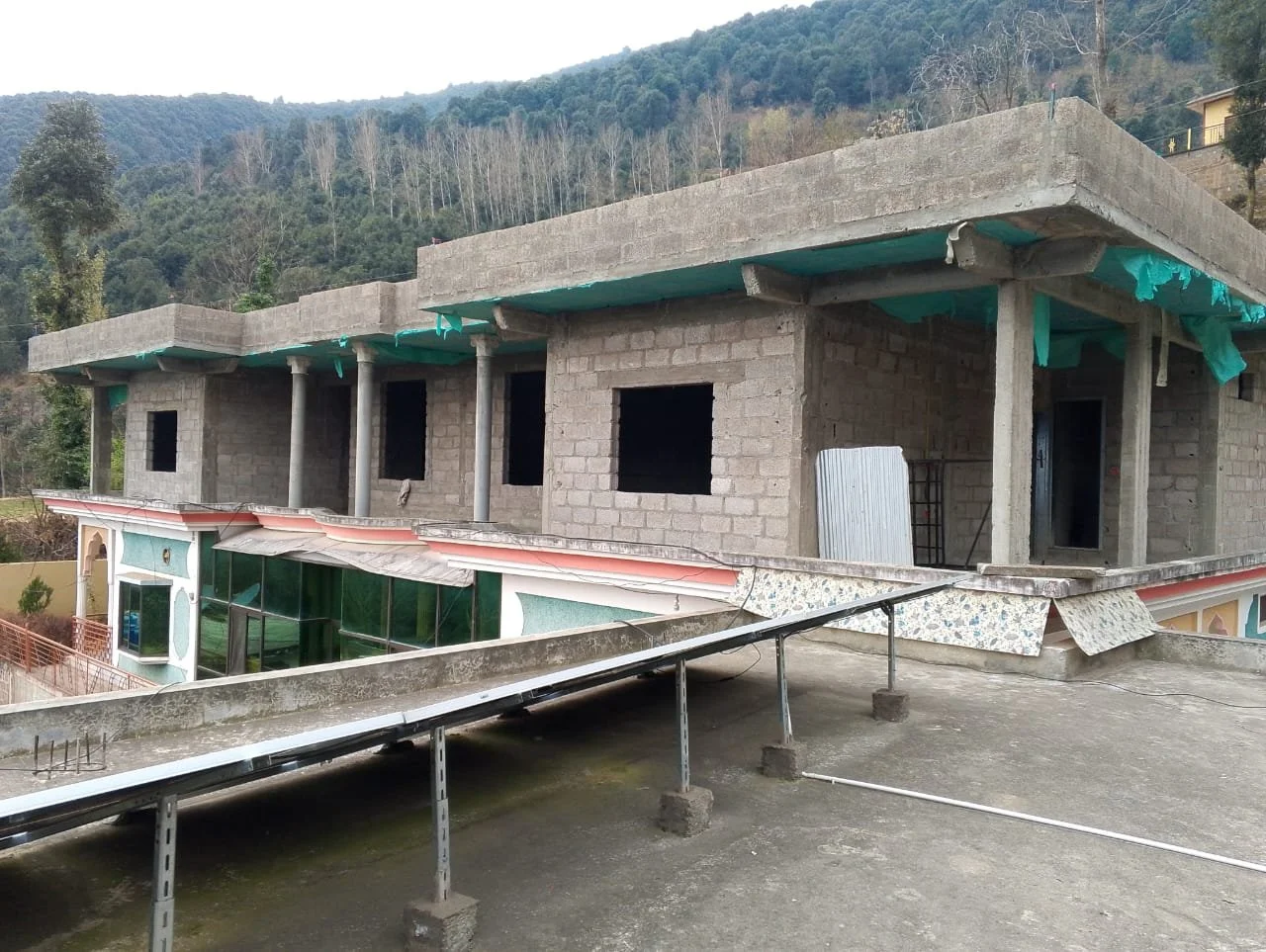 A two-story building under construction with unfinished brick walls and several window openings, situated in a hilly area with trees on the background. The ground level has a decorative facade with large windows and a door, while the upper level has 