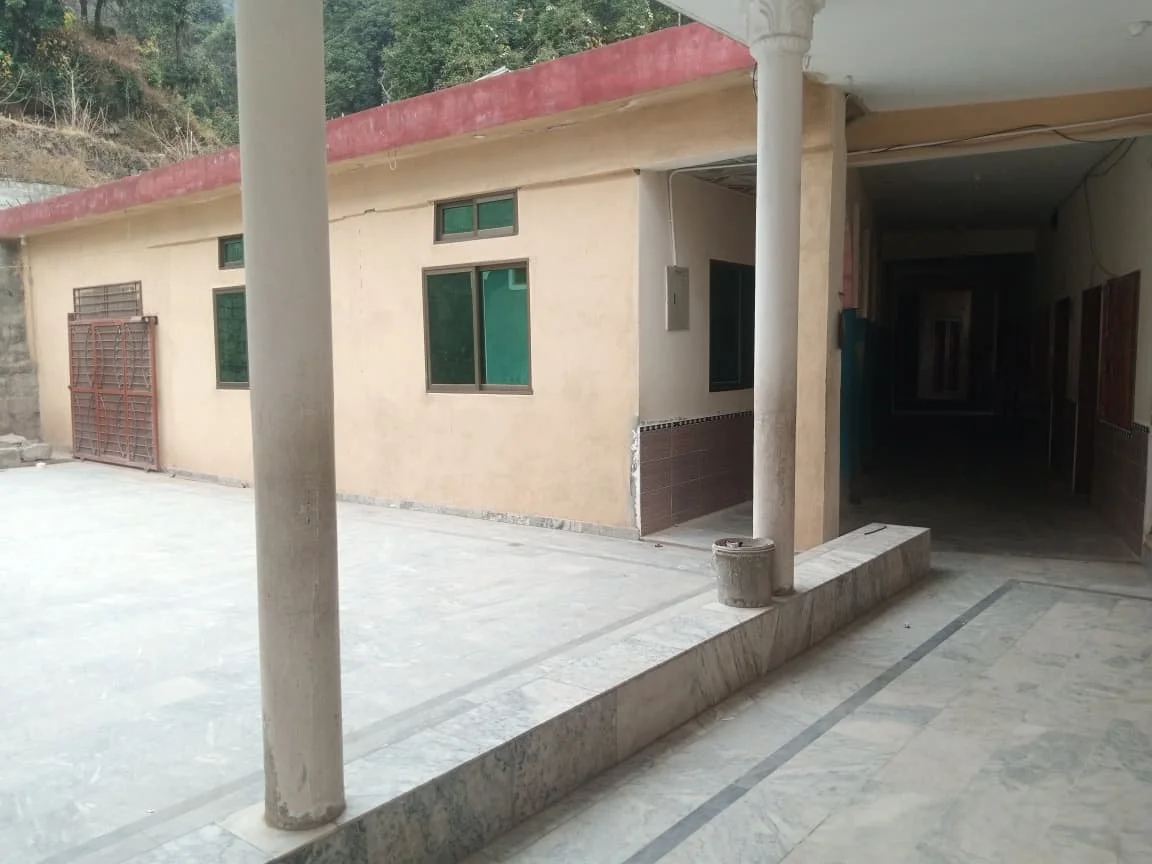 Exterior view of a beige building with multiple green windows, a red roof, and support columns, with a small patio area and surrounding landscape.