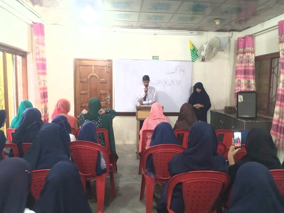 A classroom with students sitting on red chairs facing a whiteboard. A young boy in a white shirt is speaking at a podium, and a woman in black clothing stands beside him. The classroom has pink curtains, a flag, a ceiling fan, and a speaker.