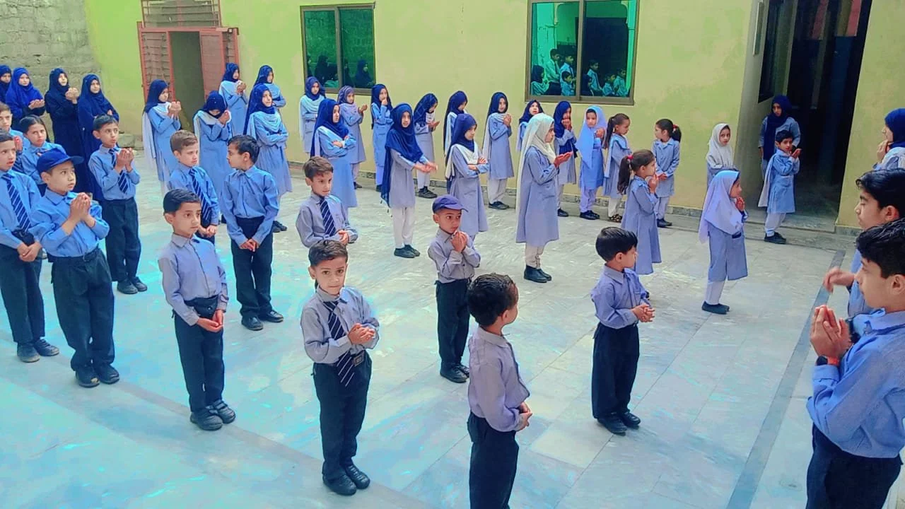School children dressed in blue uniforms standing in formation in a courtyard, with some students wearing headscarves and praying.