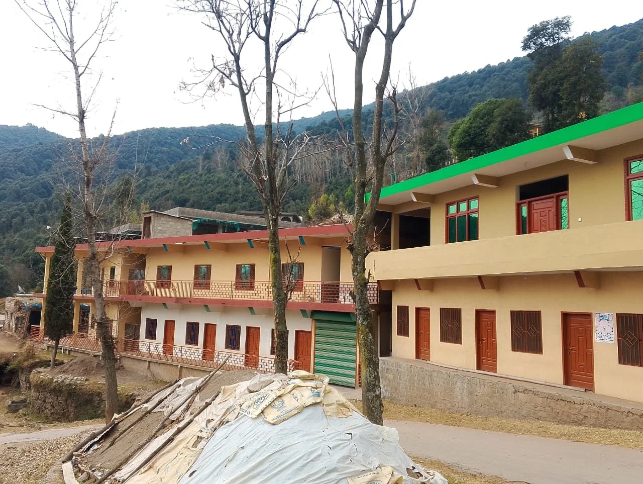 Two buildings under construction in a rural area with mountain hills in the background. Bare trees are in front, and construction debris is in the foreground.