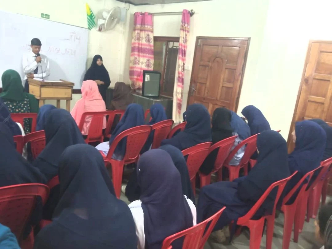 Students attending a classroom lecture with a male student speaking at the front, a woman in black seated behind, and female students wearing hijabs sitting on red chairs facing the teacher.
