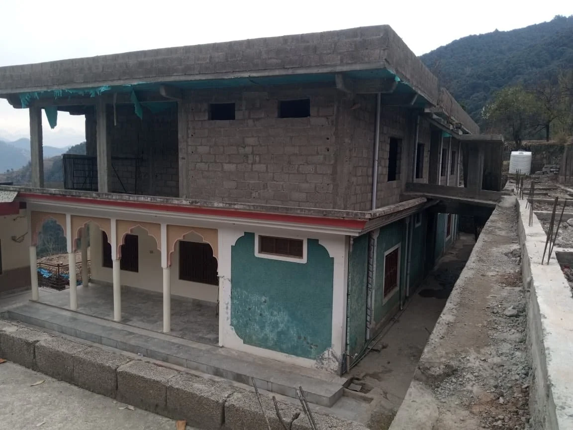 A two-story building under construction with a painted lower floor and exposed concrete upper floor, situated on a hillside with mountainous terrain in the background.