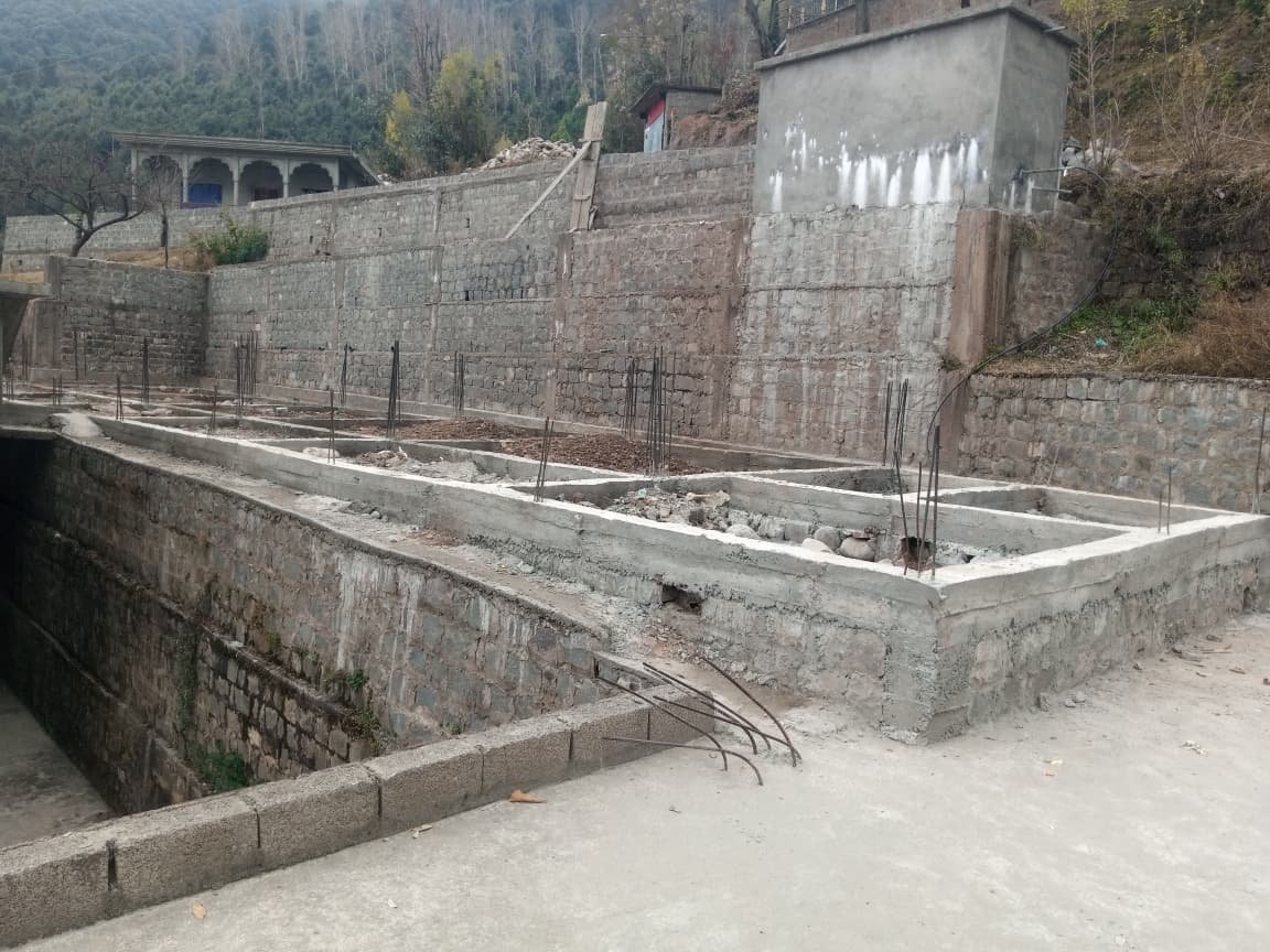 Construction site with concrete foundations and rebar for a building, set against a hillside with stone retaining walls and small structures in the background.