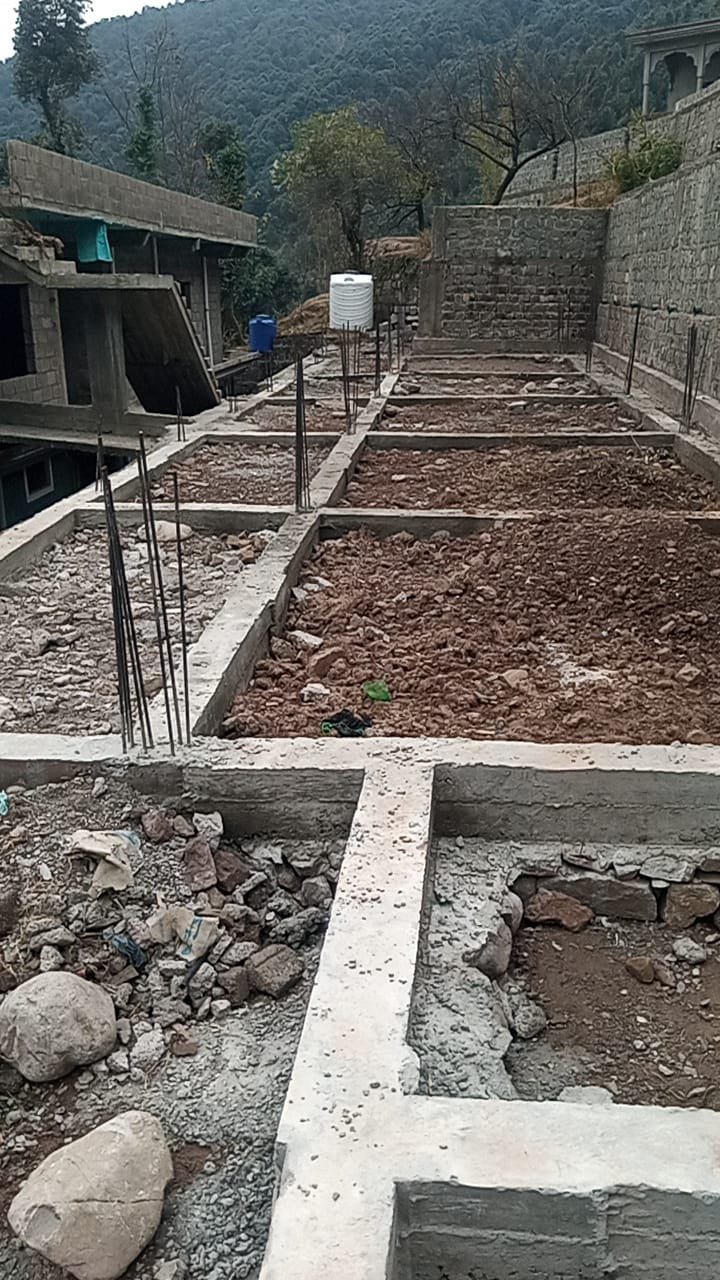 Construction site with concrete foundation and framing for building columns, surrounded by rocky terrain and hillside in the background.