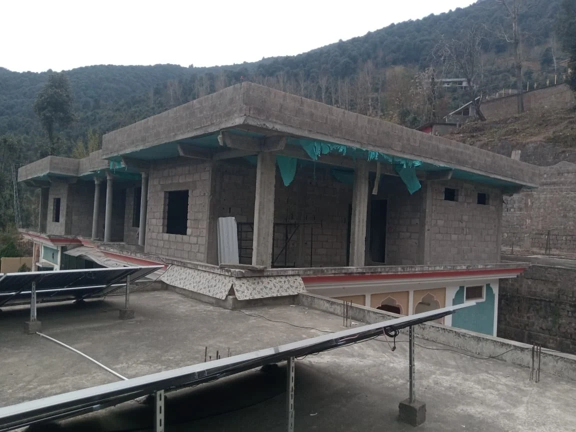 A building under construction on a flat rooftop, with solar panels installed on the roof, and a mountainous terrain in the background.