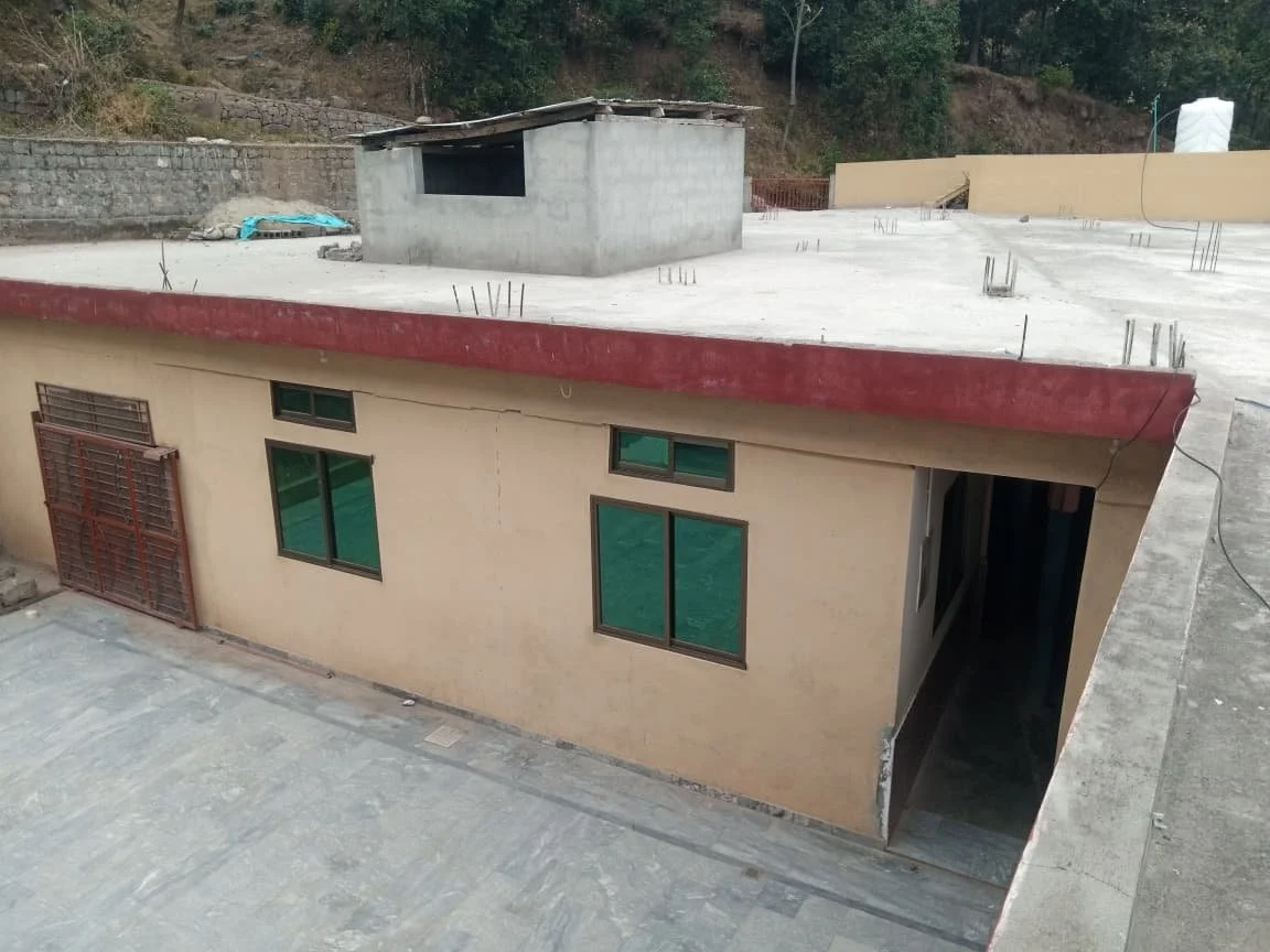 Construction site of a building with beige walls, green windows, and a flat concrete roof with exposed steel rebar, on a hilly area with trees in the background.