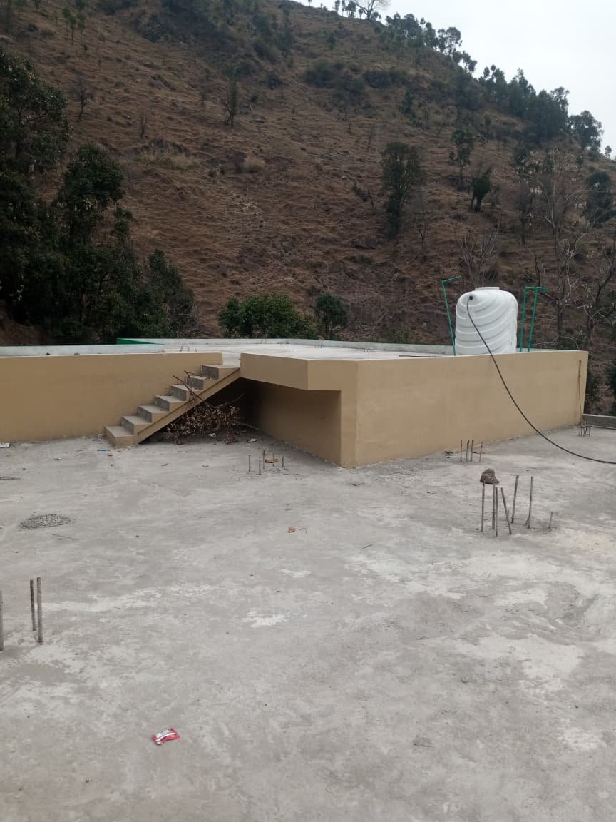 Construction site with a small staircase, a beige wall, and a white water tank on a hill with sparse trees in the background.