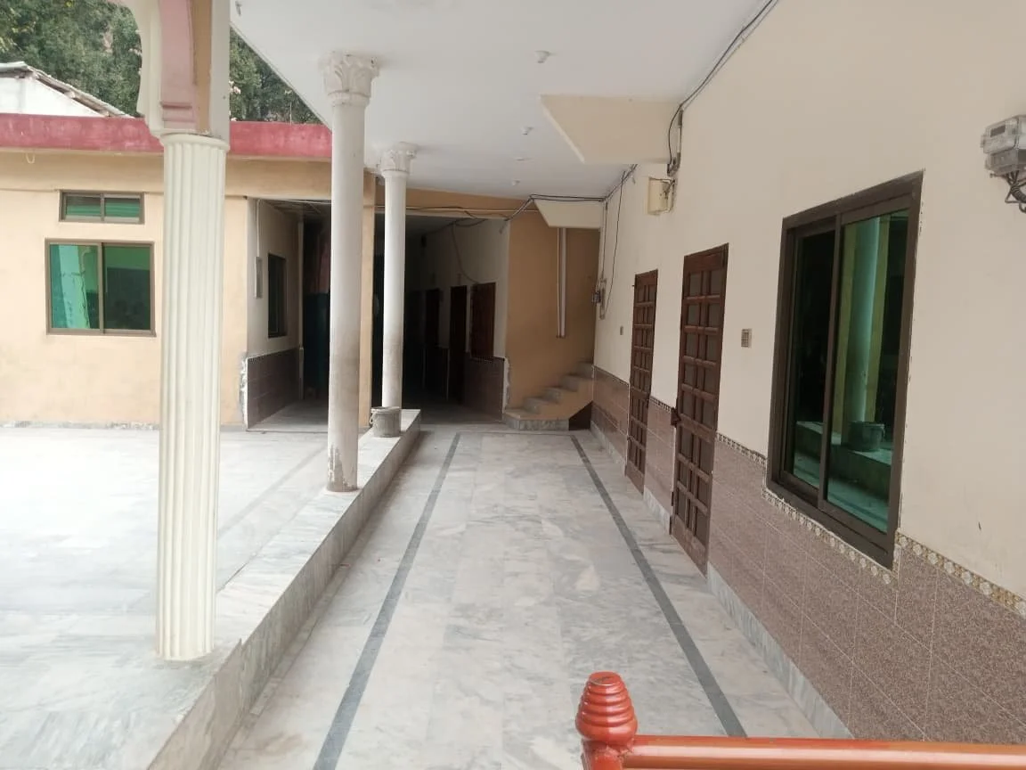 Outdoor corridor with marble flooring, white columns, and windows with wooden frames on the right, leading to a shaded area with stairs in the background.