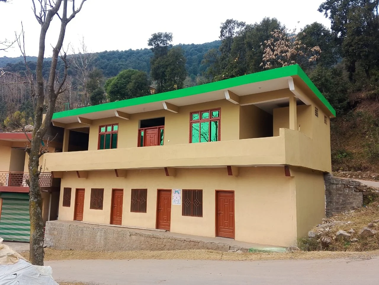A two-story house with a green roof, yellow walls, red doors, and windows, situated on a slightly elevated ground with a mountain and trees in the background.