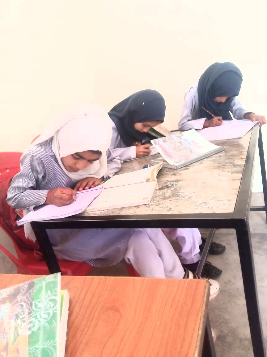 Three young girls wearing headscarves sitting at a table studying and writing in notebooks.