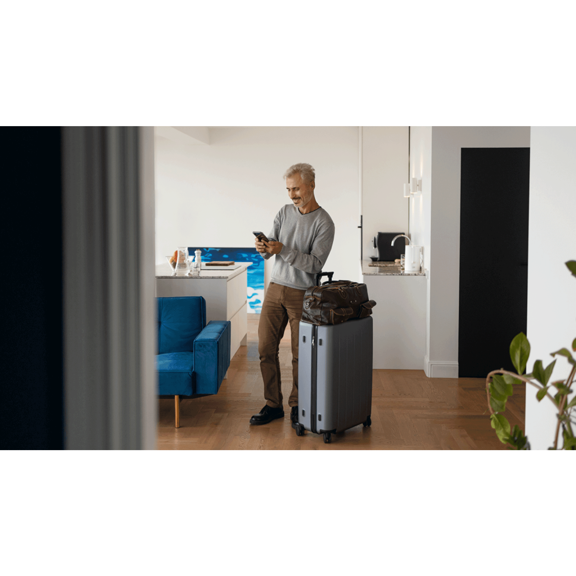 A smiling man with gray hair standing in a modern hotel room, looking at his phone. He has a suitcase and a brown bag on top of it, and is dressed casually in a gray sweater and brown pants.