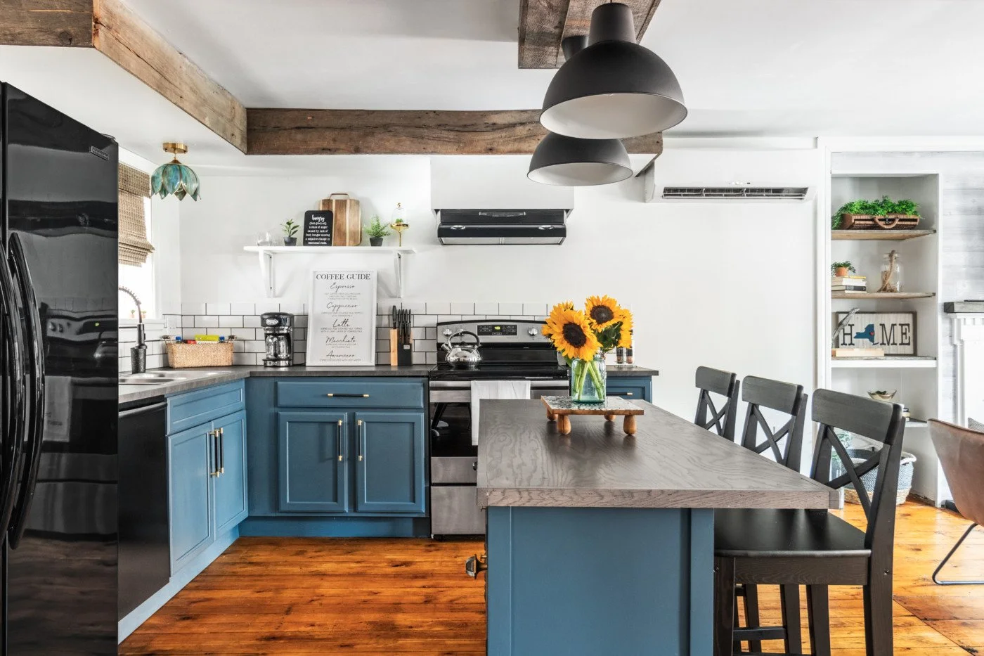A modern kitchen with blue cabinets, a wooden dining table with black chairs, and sunflower centerpiece. Features include white subway tile backsplash, black appliances, hanging black and white pendant lights, and decorative shelves with plants and books.