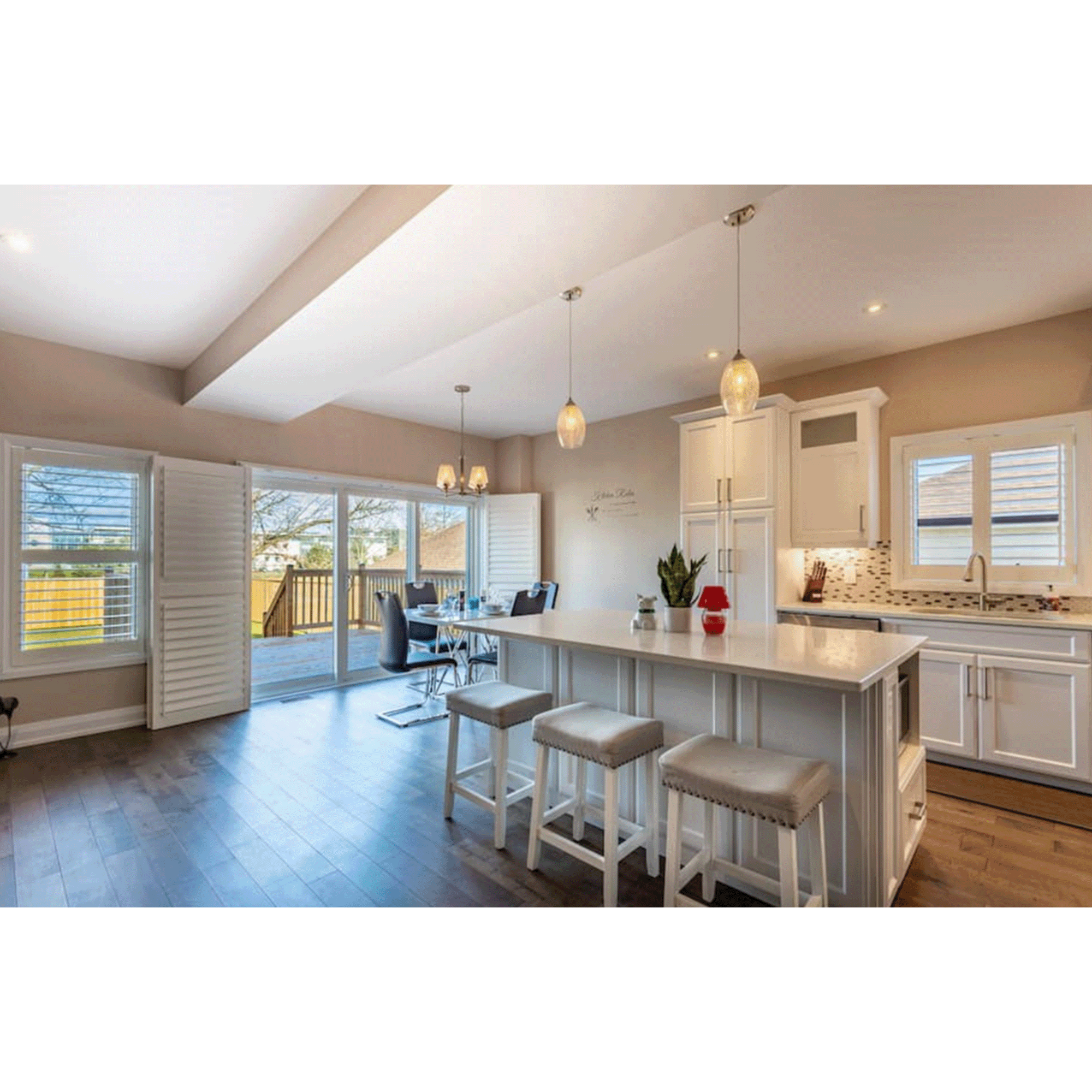 Modern kitchen and dining area with white cabinets, a kitchen island, and wooden flooring, sunlight streaming through large windows and sliding glass door leading to outdoor deck.