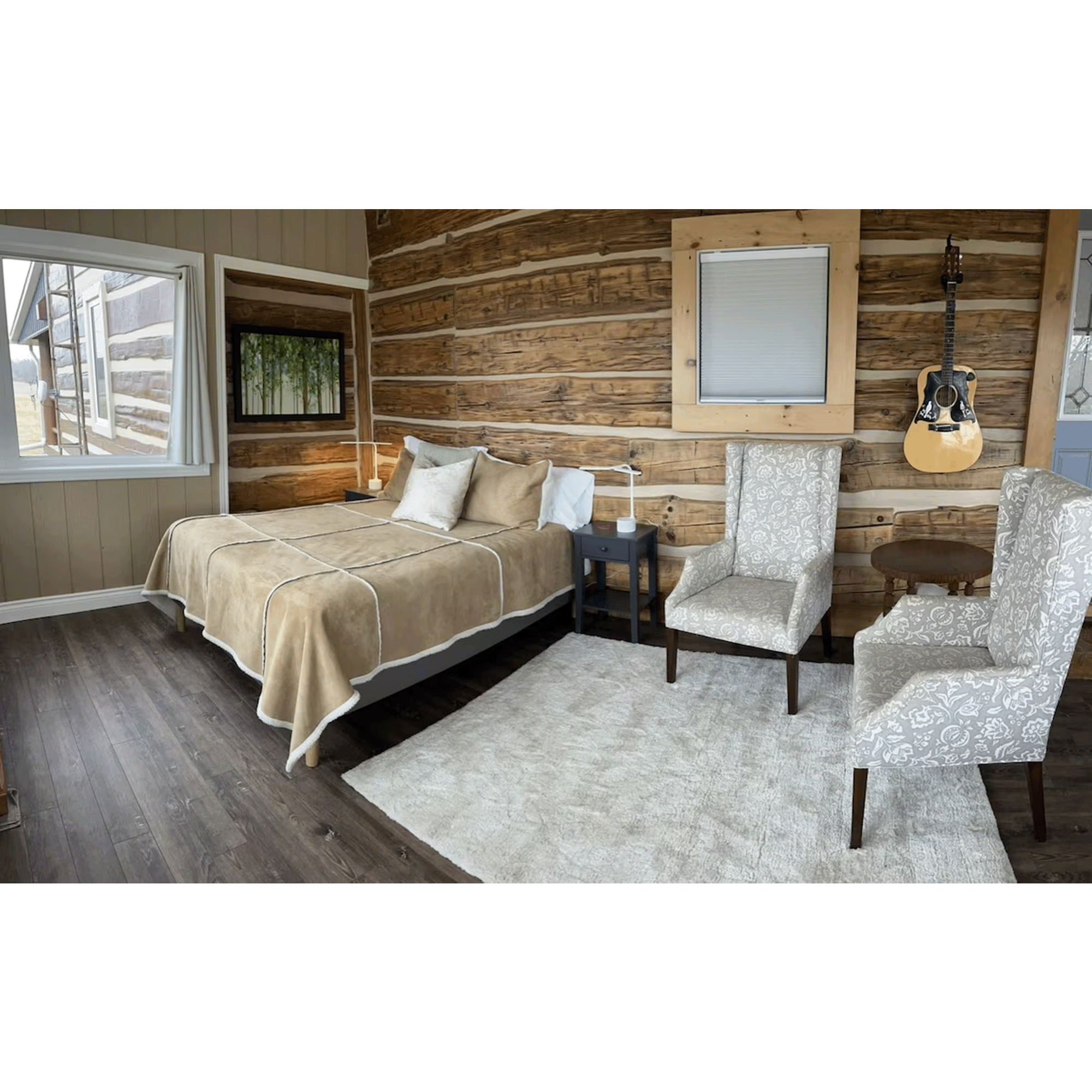 Cozy bedroom with wooden log walls, a bed with beige bedding and pillows, a black nightstand, two patterned armchairs, a small wooden table, a guitar hanging on the wall, and windows showing an outdoor view.