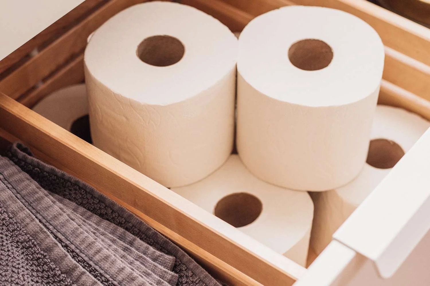 Six rolls of white toilet paper stacked in a wooden crate, with a piece of gray textured towel visible in the foreground.