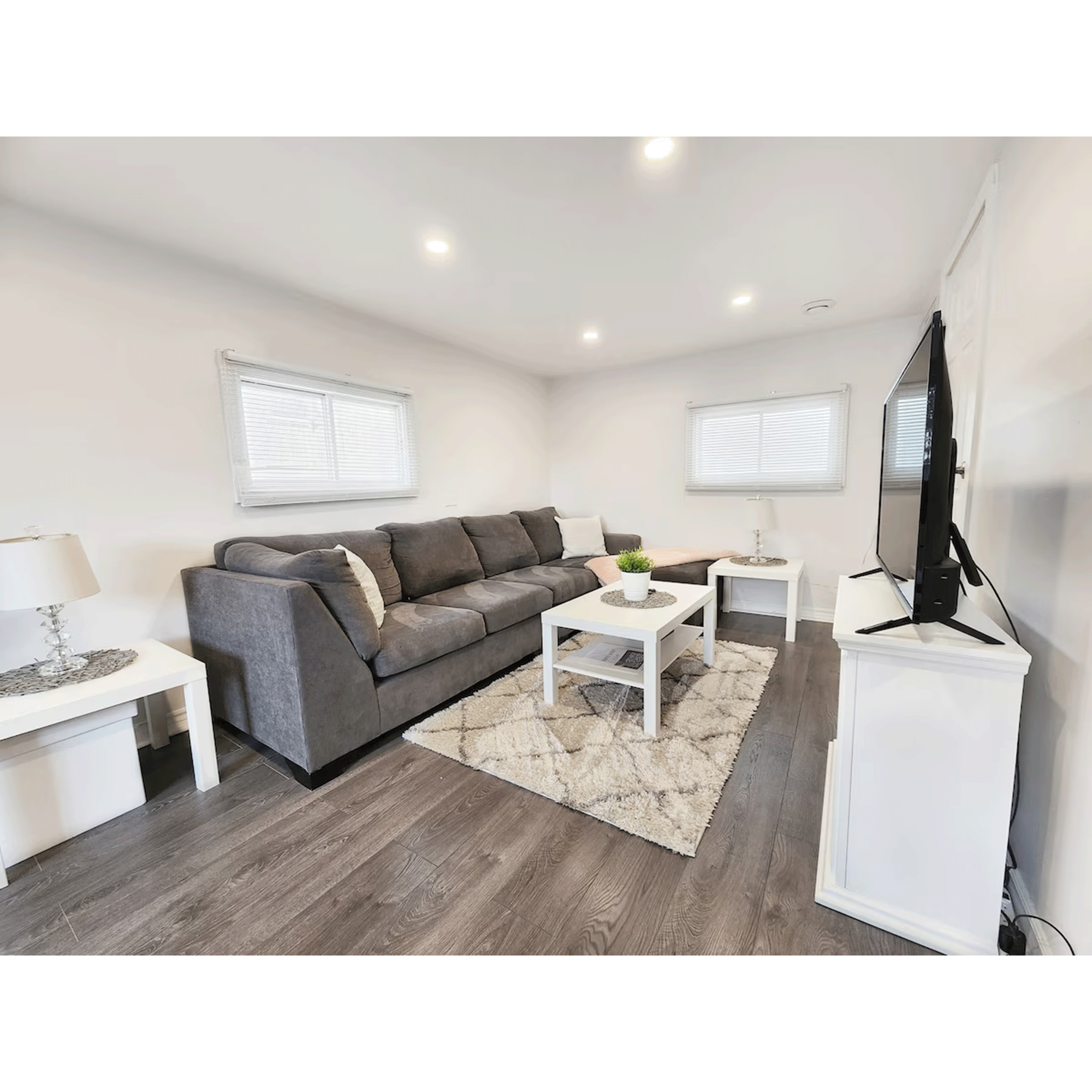 Bright living room with white walls, dark hardwood floor, gray sofa, white coffee table with plant, patterned rug, side tables with lamps, and a large flat-screen TV on white stand.