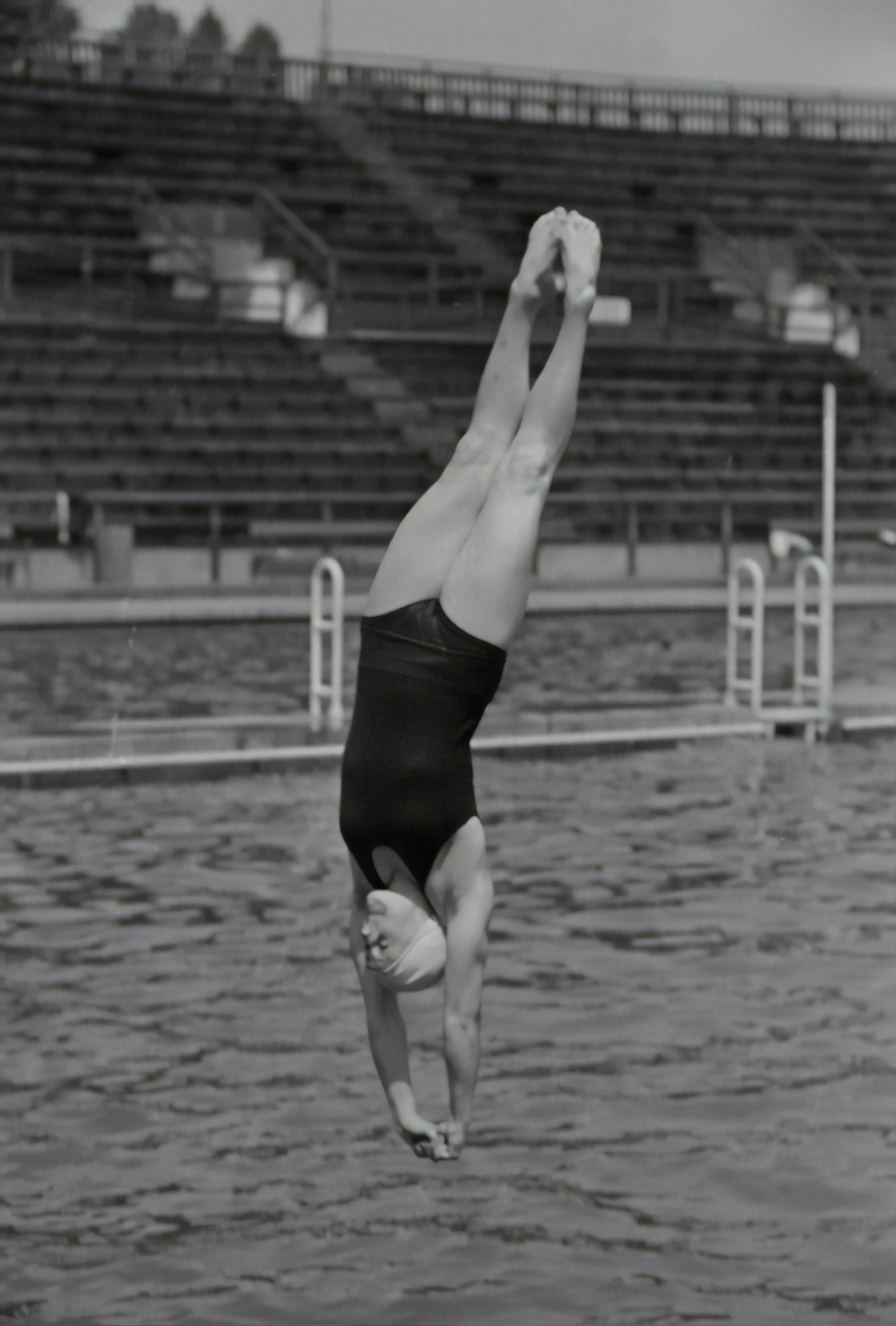 A woman in a black swimsuit is performing a handstand on water in an outdoor pool with empty bleachers in the background.