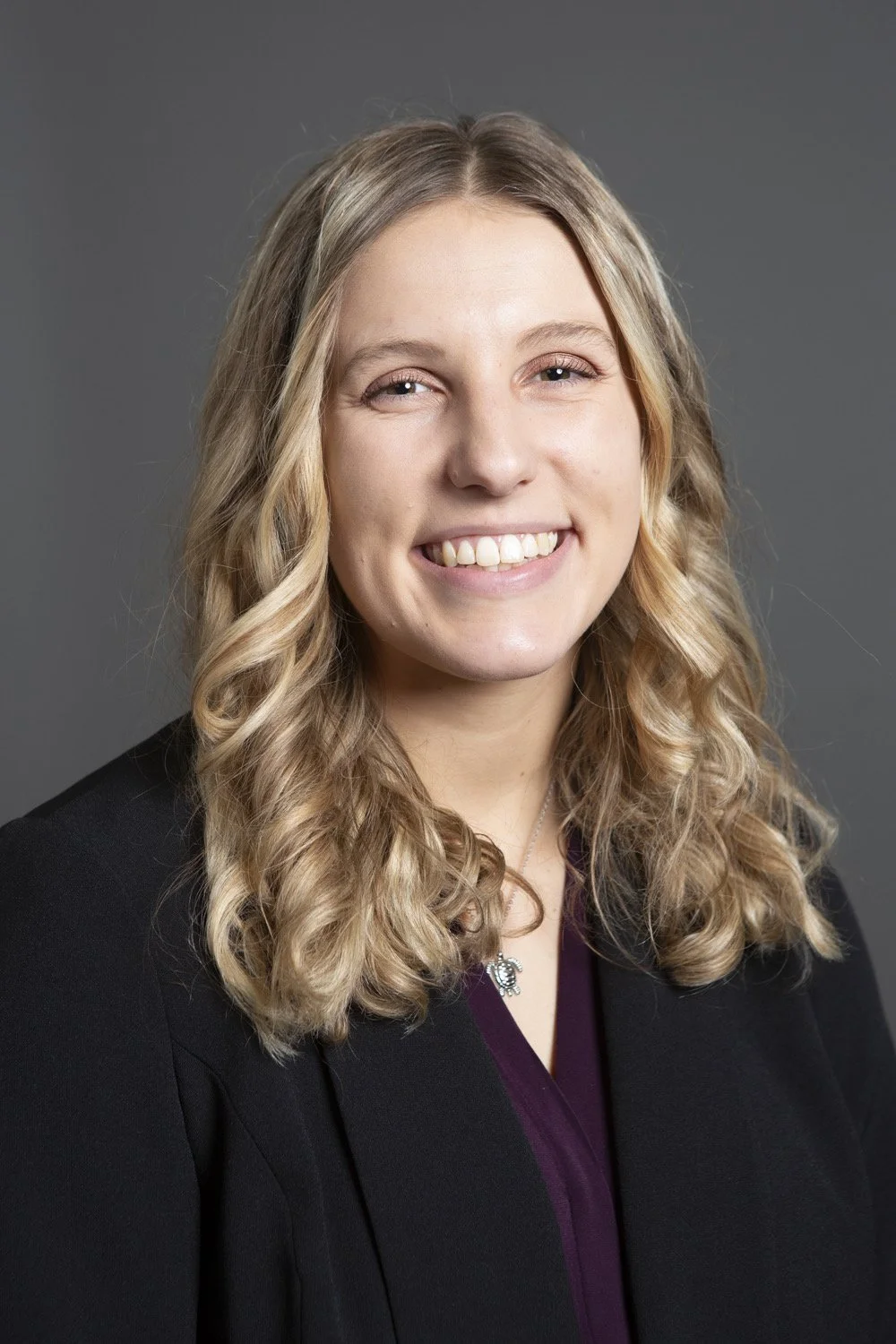 Portrait of a woman with blonde curly hair, smiling, wearing a black blazer and a purple top, against a gray background.