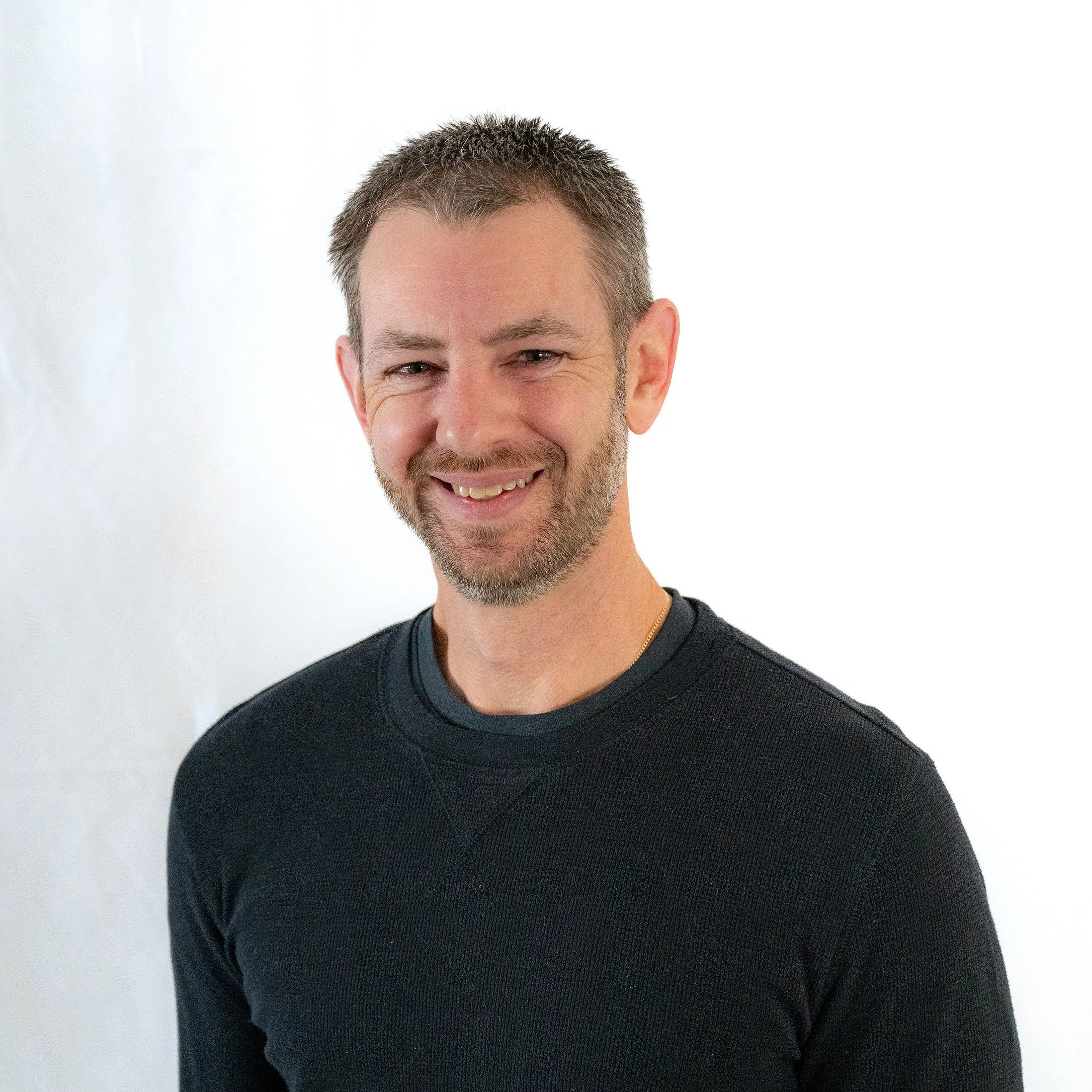 A smiling man with short dark hair, beard, and wearing a black shirt, standing against a white background.