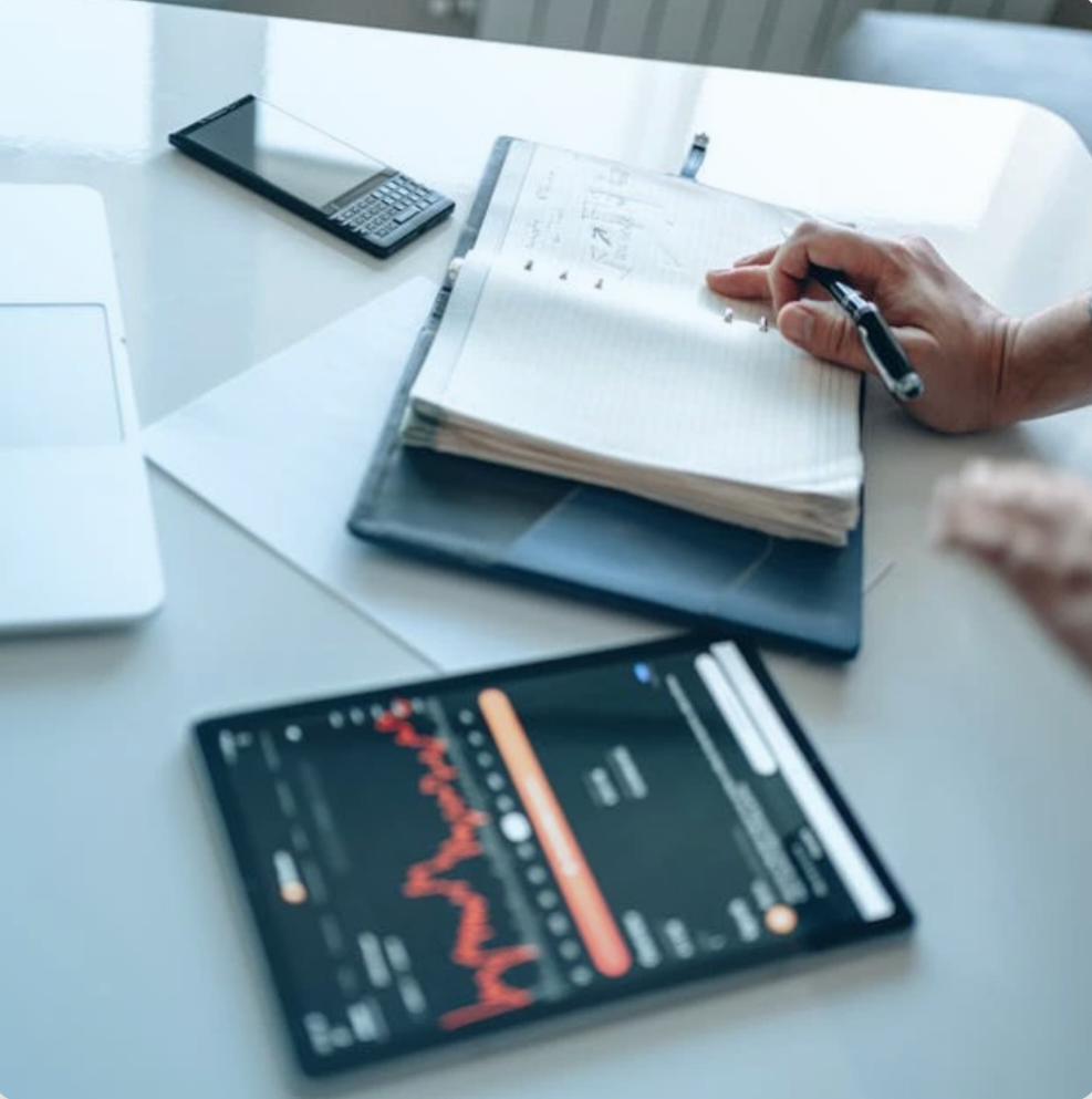 Una scrivania con un laptop, uno smartphone con grafici finanziari, diari e una mano che scrive in un taccuino.
