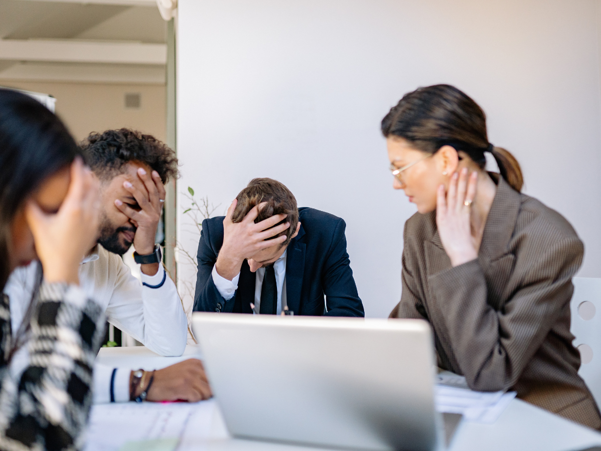 Four professionals sitting around a table in an office, each with their head in their hands, appearing stressed or overwhelmed while reviewing documents on a laptop.