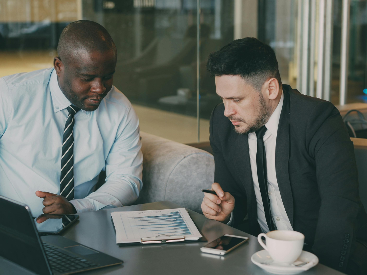 Two business professionals sitting at a table in front of a laptop reviewing printed data reports.