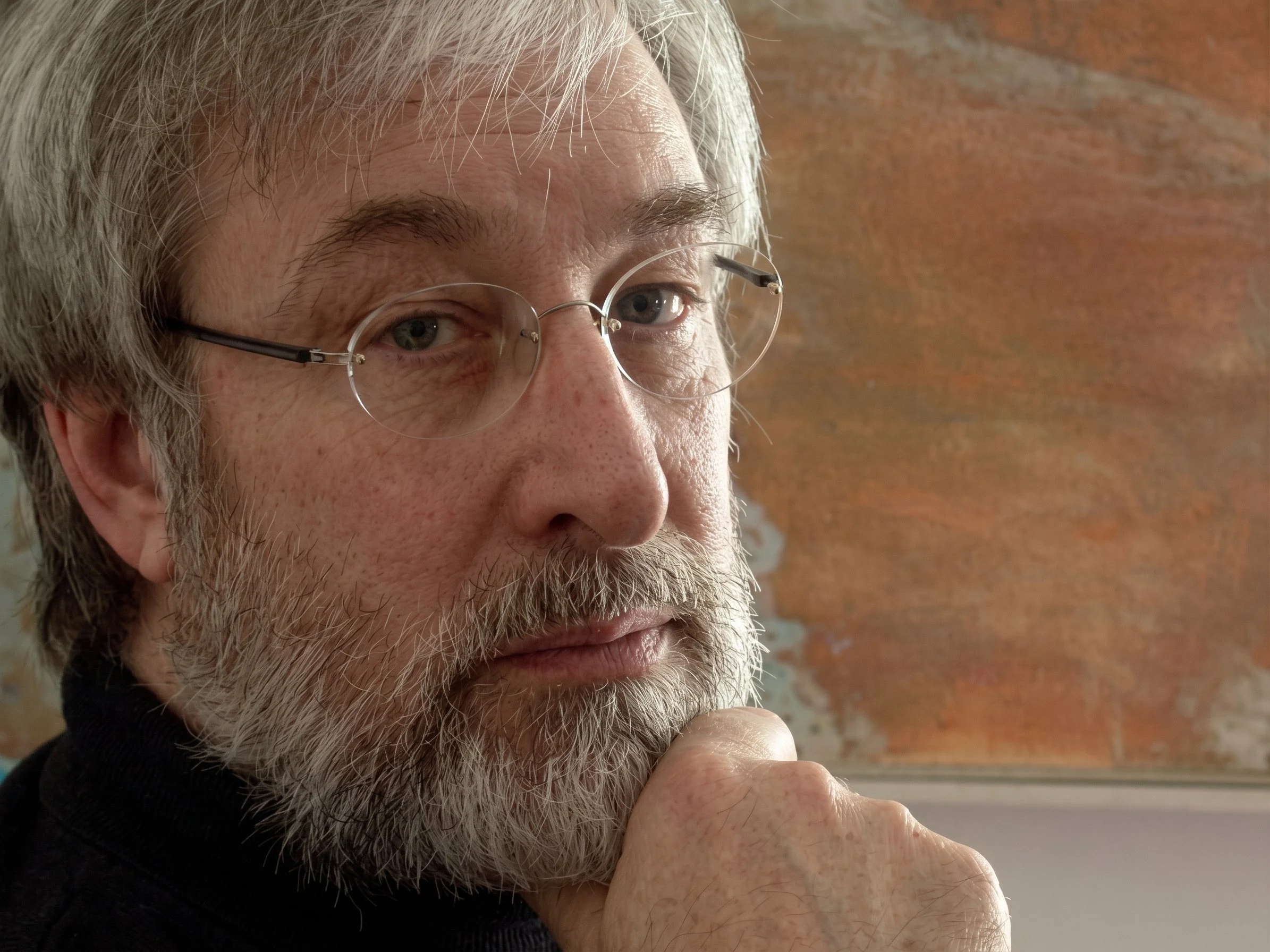 Close-up of an older man with gray hair and beard, wearing glasses, thinking with a hand on his chin, against a brick wall background.