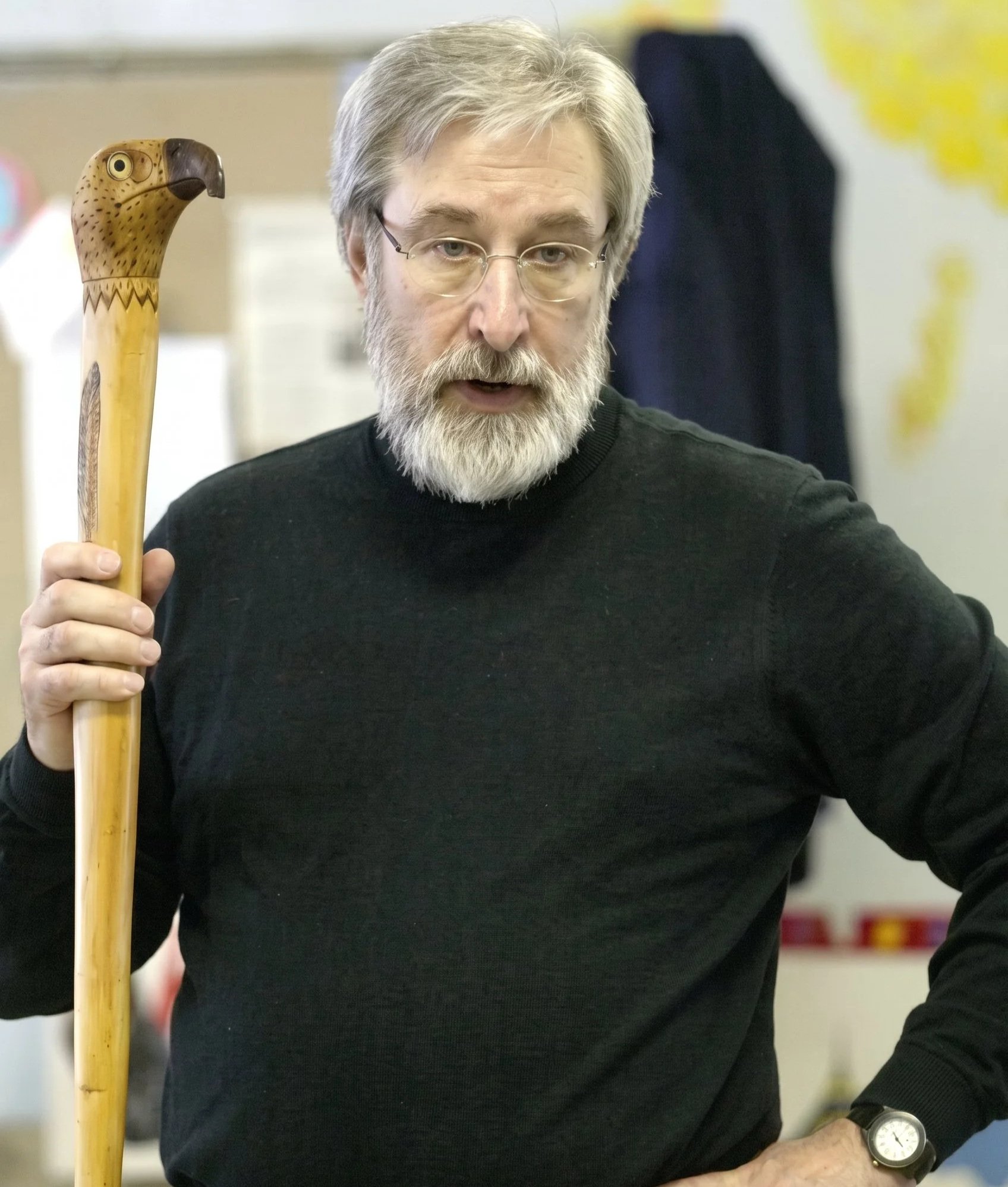 A man with glasses and a gray beard holding a carved wooden walking stick, wearing a black shirt, with a wristwatch, in an indoor setting with a blurred background that includes a black jacket hanging and yellow decorations.