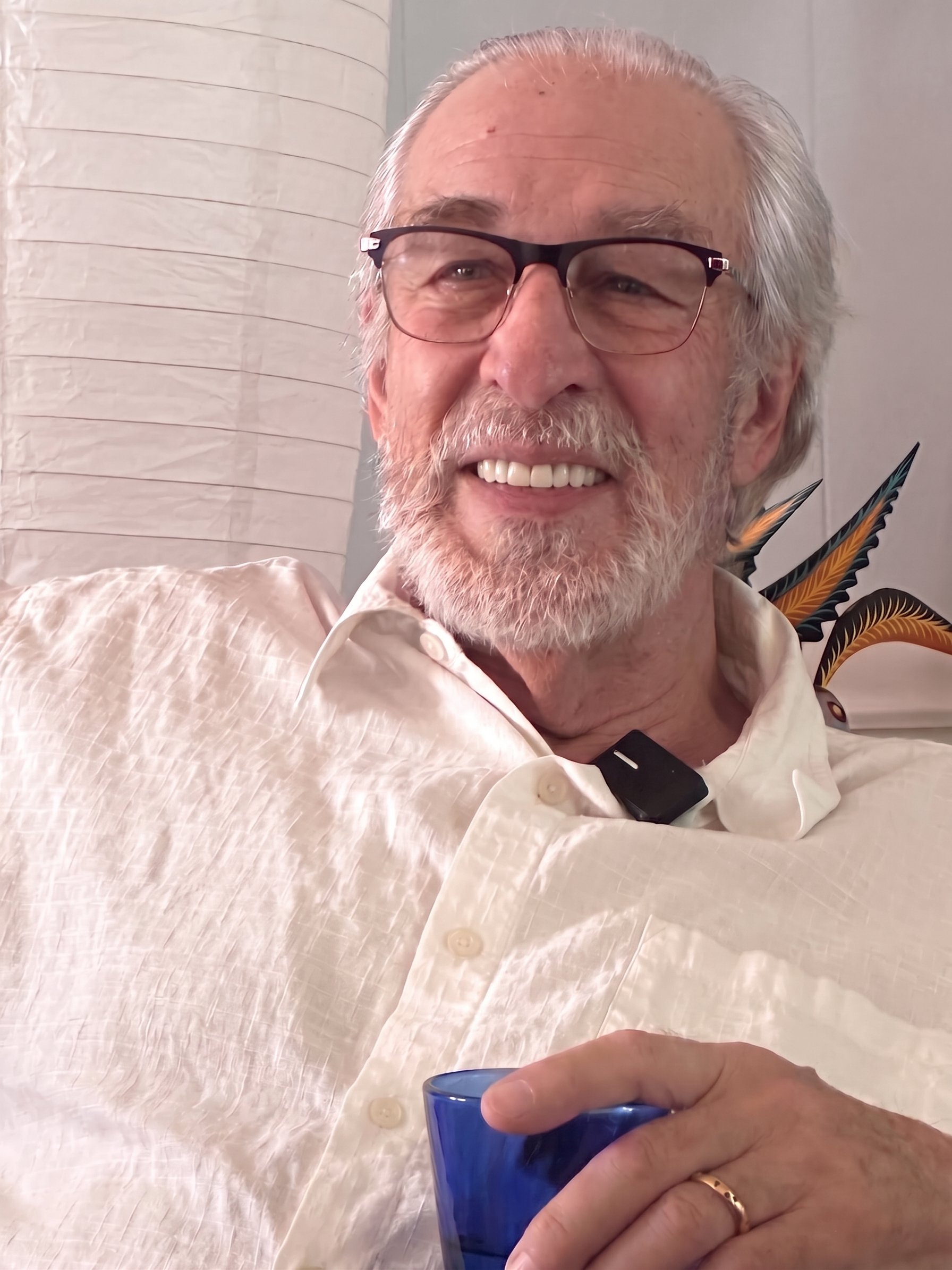 An older man with white hair, glasses, and a beard is smiling. He is wearing a cream-colored shirt and holding a blue glass. A hand with a wedding ring holds the glass. In the background, part of a decorative plant and a light-colored lamp are visible.
