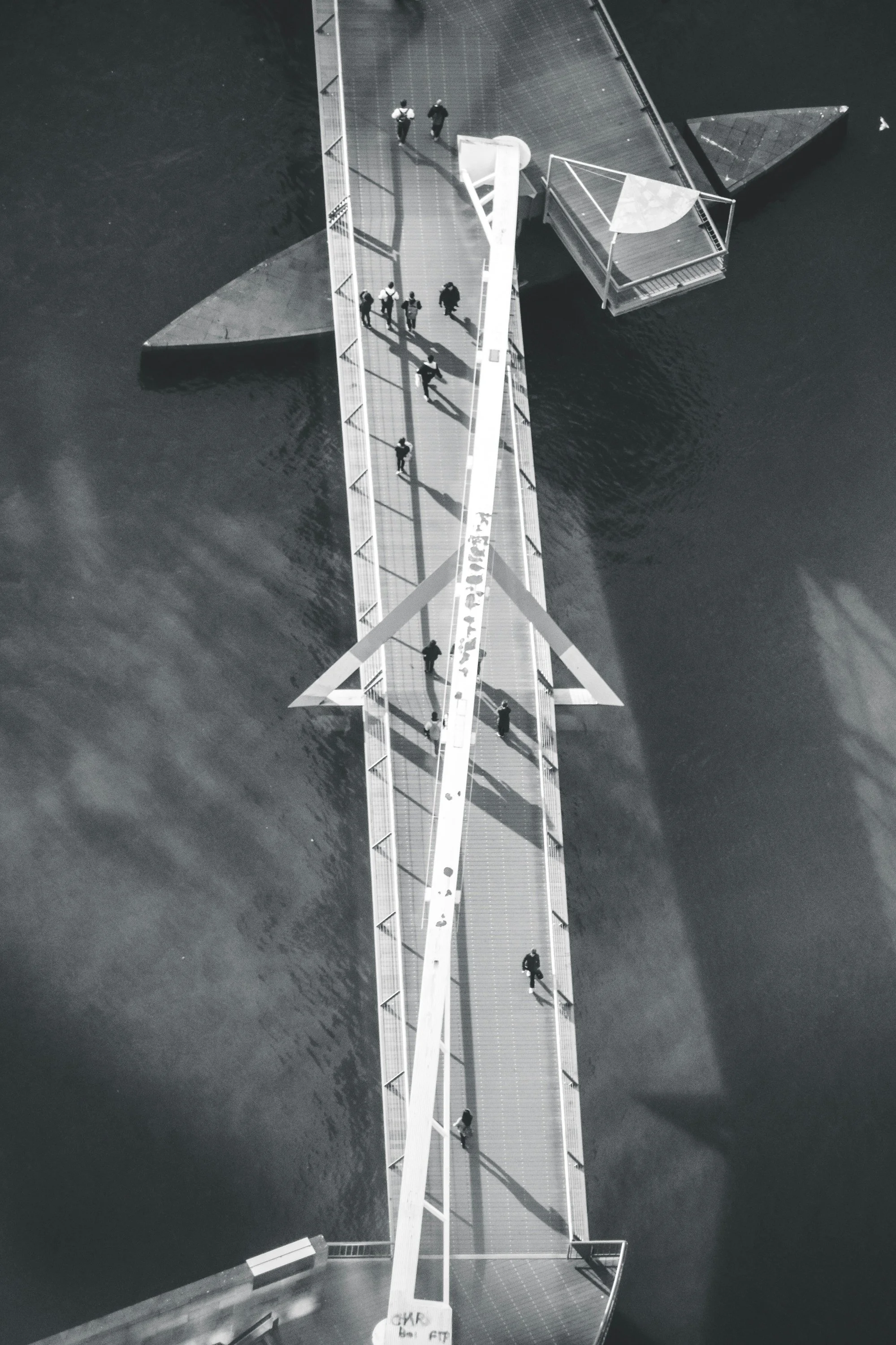 Aerial view of a modern, white, diagonal footbridge over water with people walking on it.